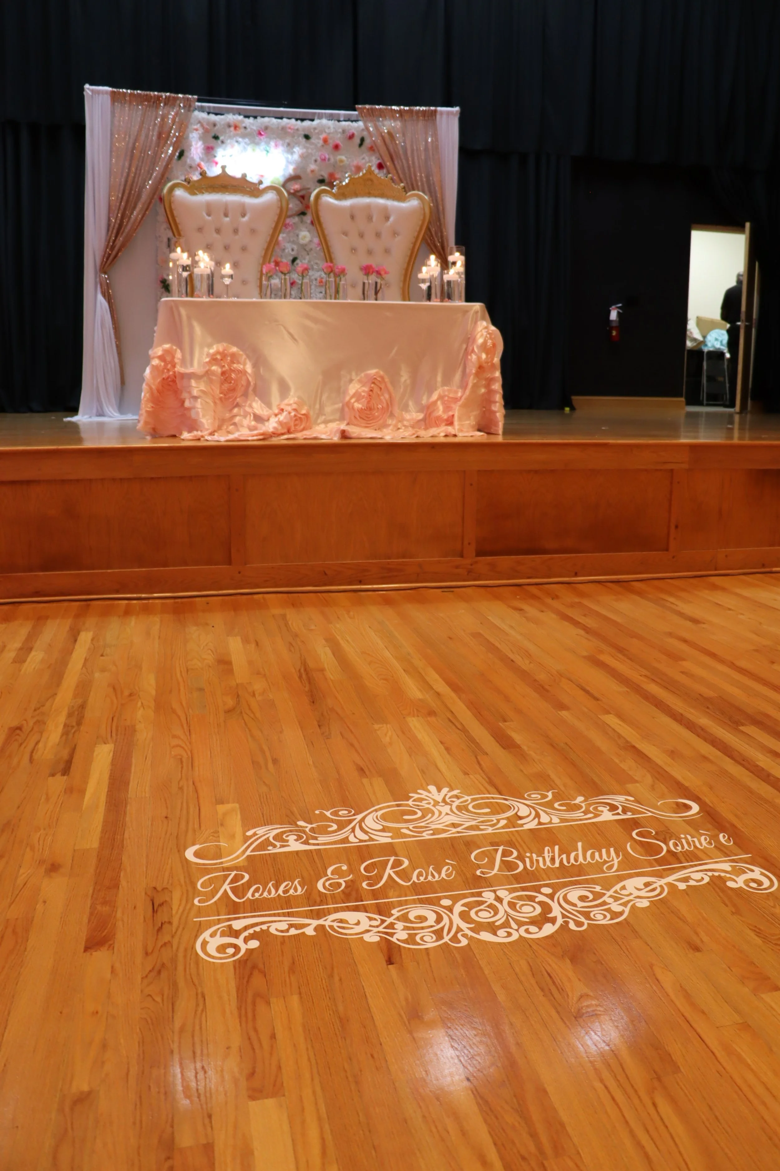 A decorated event stage with two ornate chairs, a table with pink ruffled fabric, candles, and pink flowers. The wooden floor has a white script message that reads 'Roses & Rosé Birthday Soiree'.