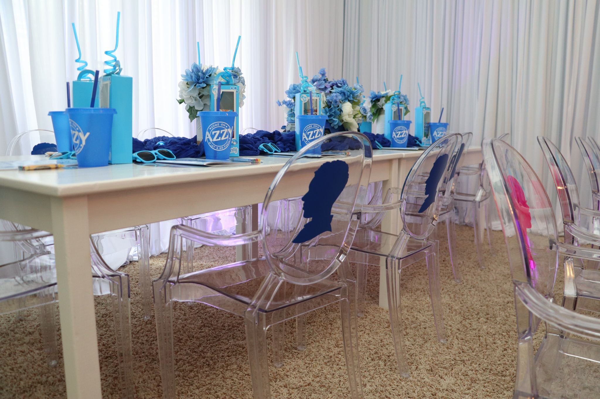 Party table decorated with blue drinks, flowers, and party favors, with transparent chairs featuring silhouette designs, set in a bright room with white curtains and beige carpet.