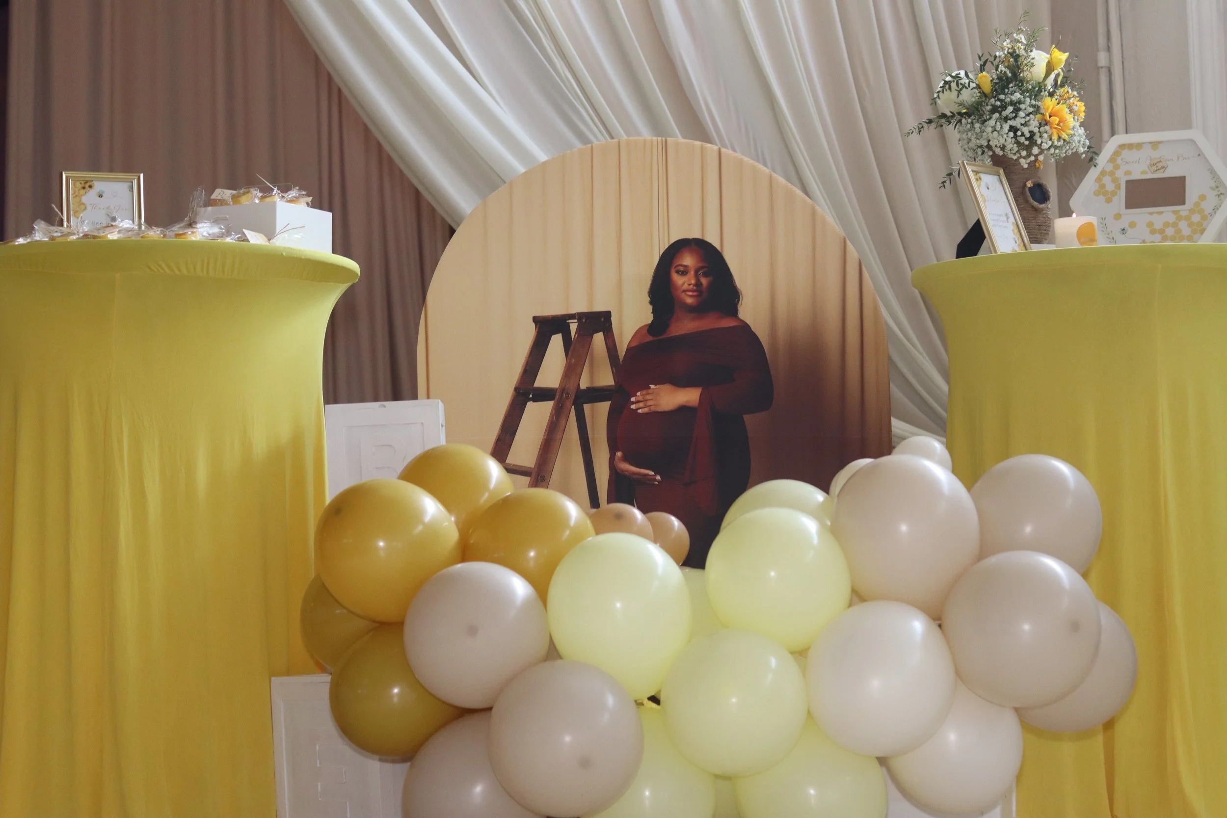 A pregnant woman in a burgundy dress standing behind a display decorated with yellow and white balloons, with yellow cloth draping and a wooden backdrop.