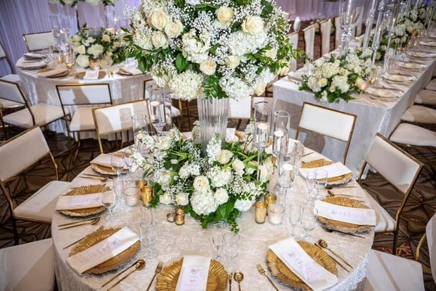 Elegant wedding reception table setting with white floral centerpiece, gold-accented plates, and glassware, arranged in a decorated banquet hall.