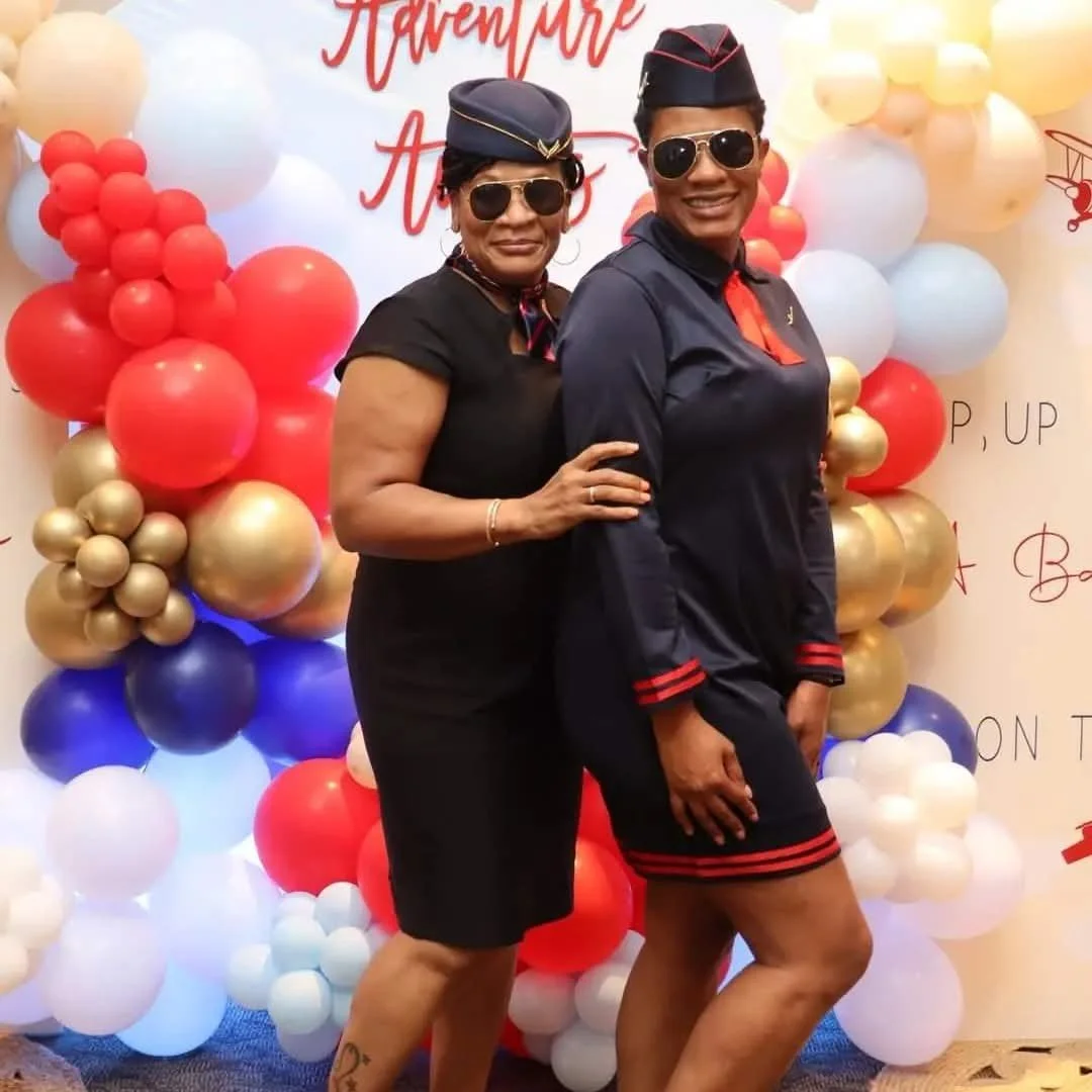 Two women dressed in pilot uniforms with sunglasses, celebrating in front of a balloon backdrop with red, gold, white, and blue balloons, and a sign possibly indicating an adventurous celebration.