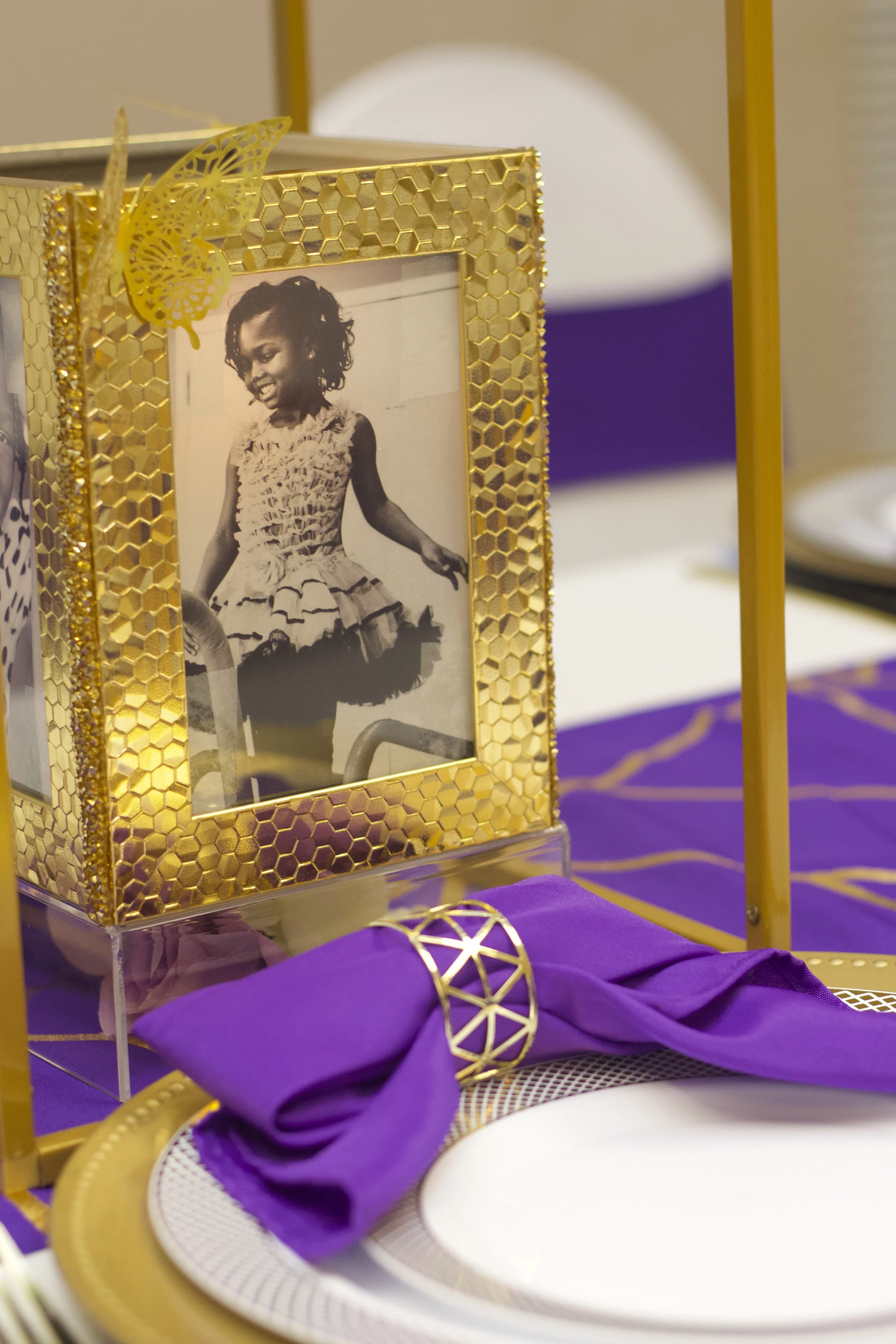 A black-and-white photograph of a young girl in a dress, displayed in a gold geometric frame on a purple tablecloth with gold accents, part of a decorated table setting.