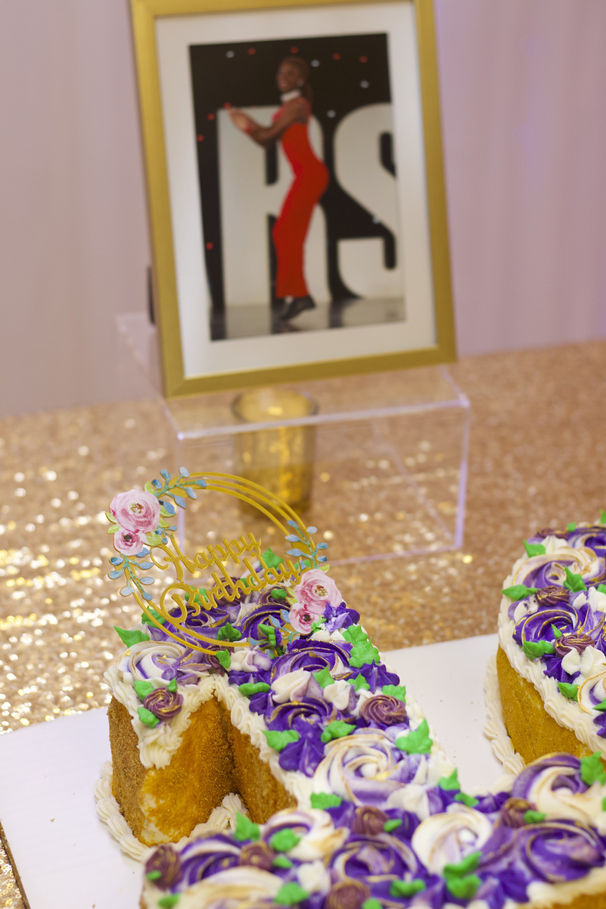 A decorated birthday cake with purple, white, and green icing flowers and a "Happy Birthday" topper; a framed photo of a woman in a red dress dancing, displayed on a gold and clear stand, in the background.