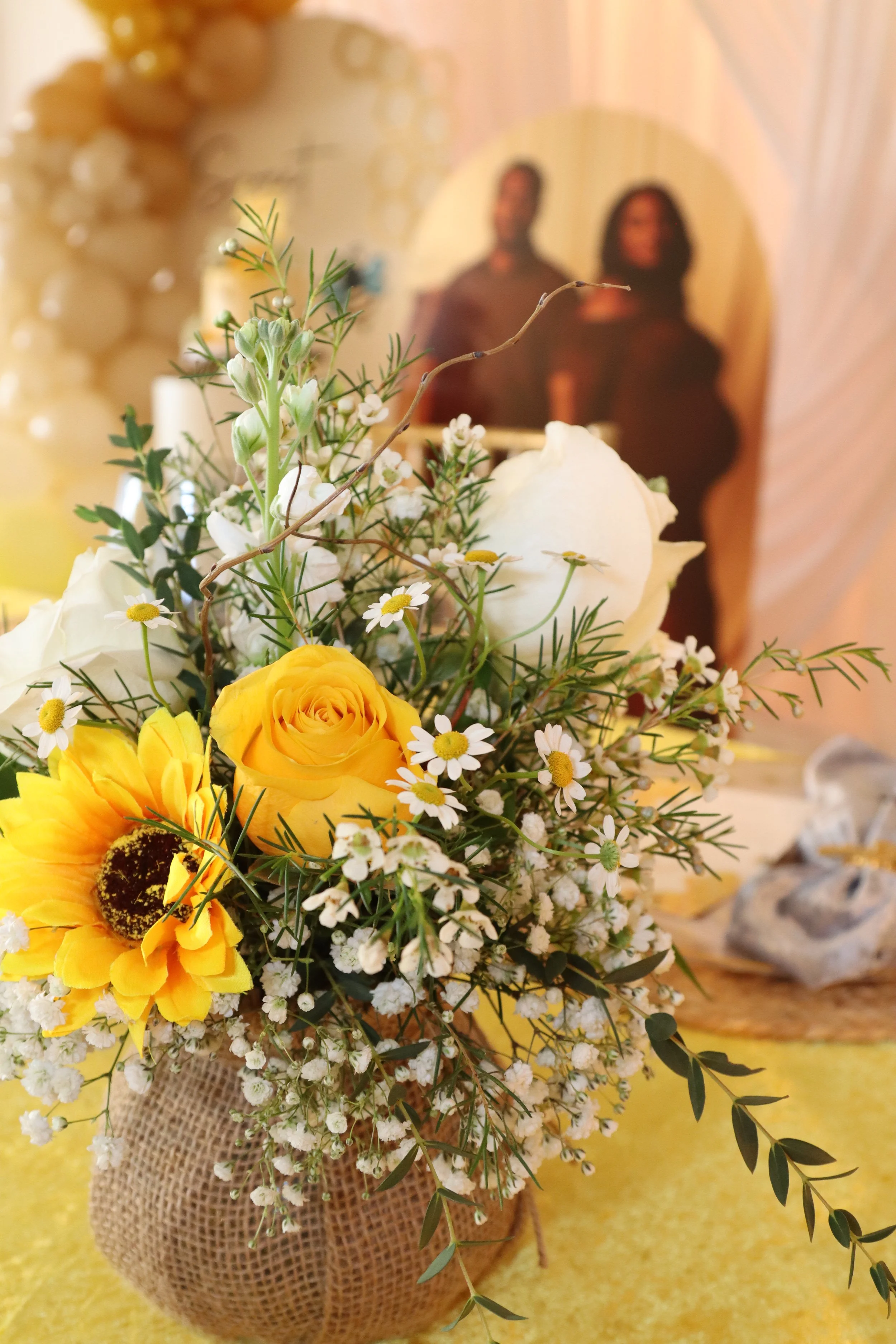 A floral arrangement featuring yellow roses, a yellow sunflower, white roses, baby's breath, and greenery in a burlap-covered vase, with a blurred photo of a couple in the background.