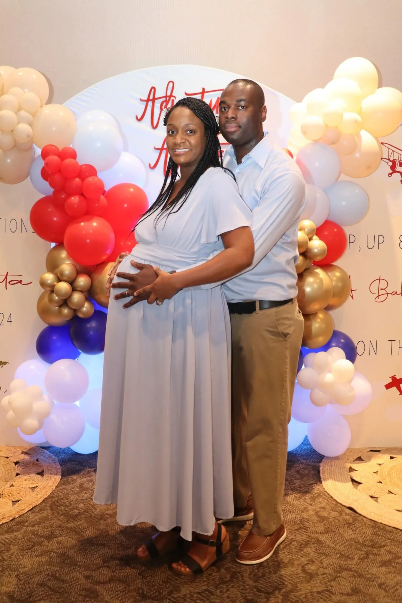 A couple, with the woman visibly pregnant, stands in front of a decorated backdrop with balloons in red, gold, white, and purple. The woman is wearing a light gray dress, and the man is wearing a light blue shirt and khaki pants. They are embracing e
