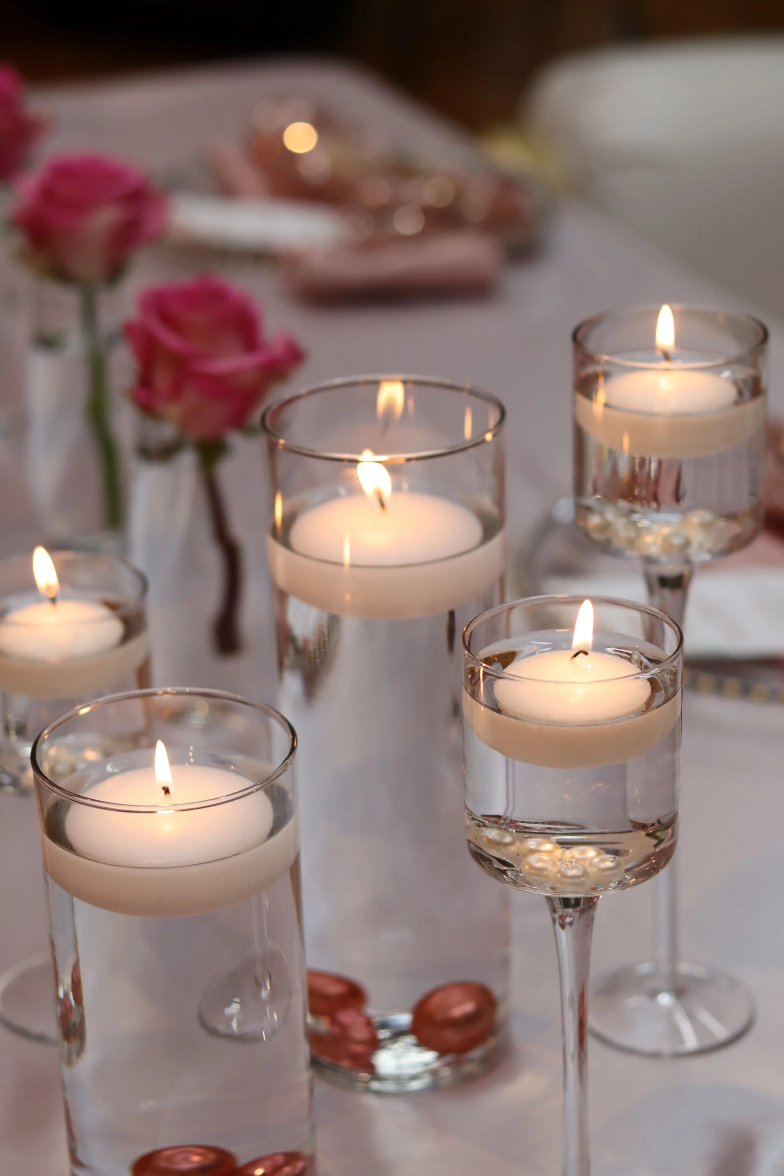 Six floating candles in clear glass holders, surrounded by pink roses on a white tablecloth.