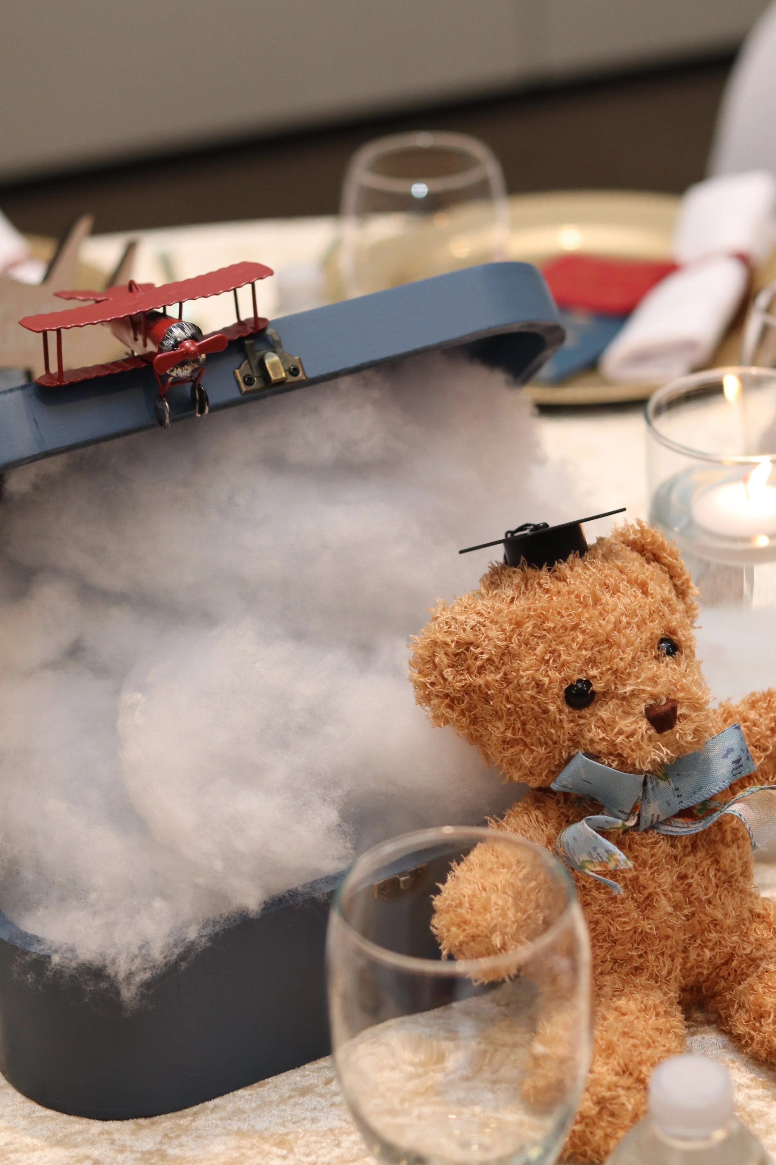 A teddy bear wearing a graduation cap and bow tie positioned in front of a decorative setup featuring a small toy airplane and a cloudy backdrop, with teacups and candles on a table.