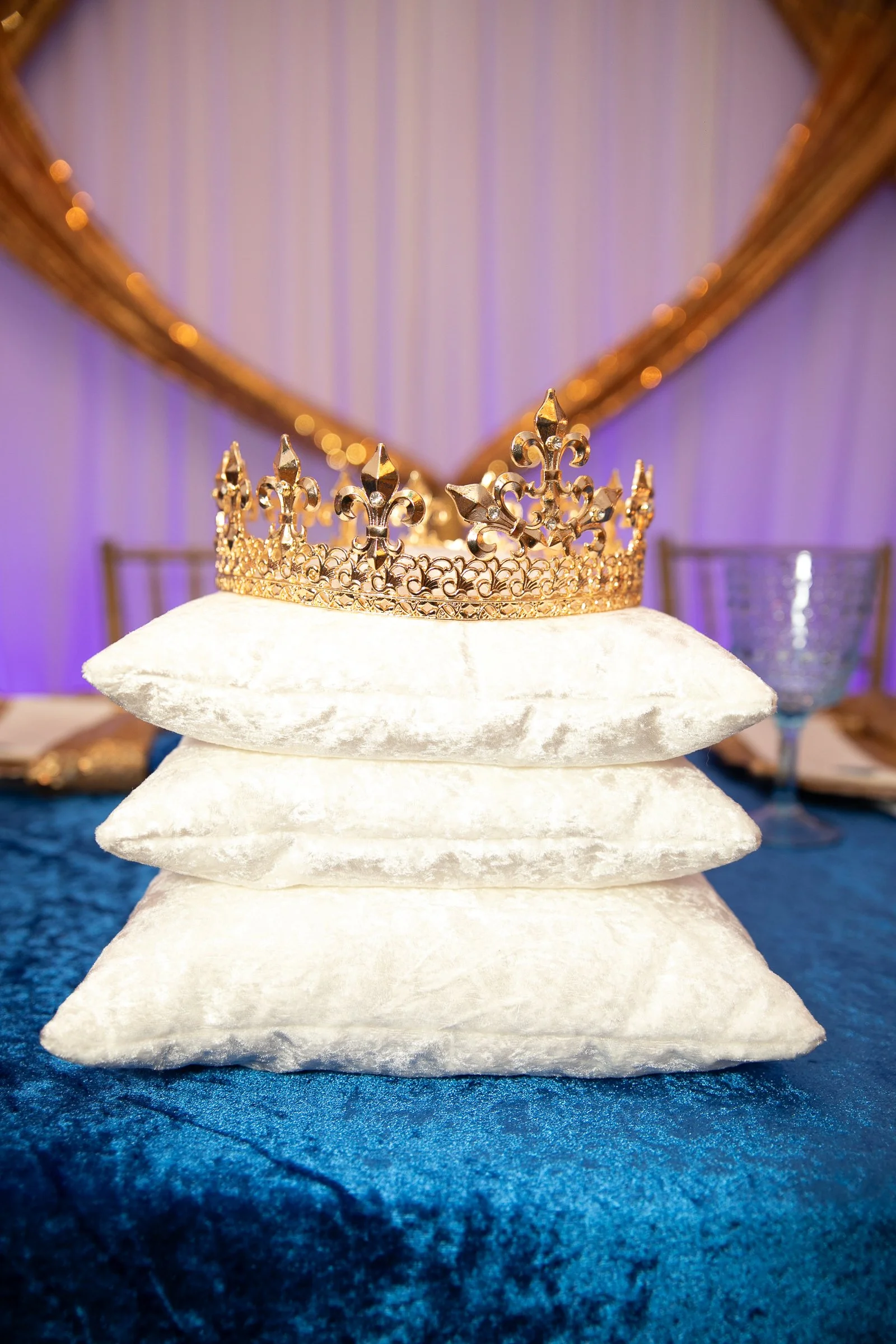 A golden crown placed on three stacked white velvet pillows on a blue tablecloth at a decorated event with purple lighting.
