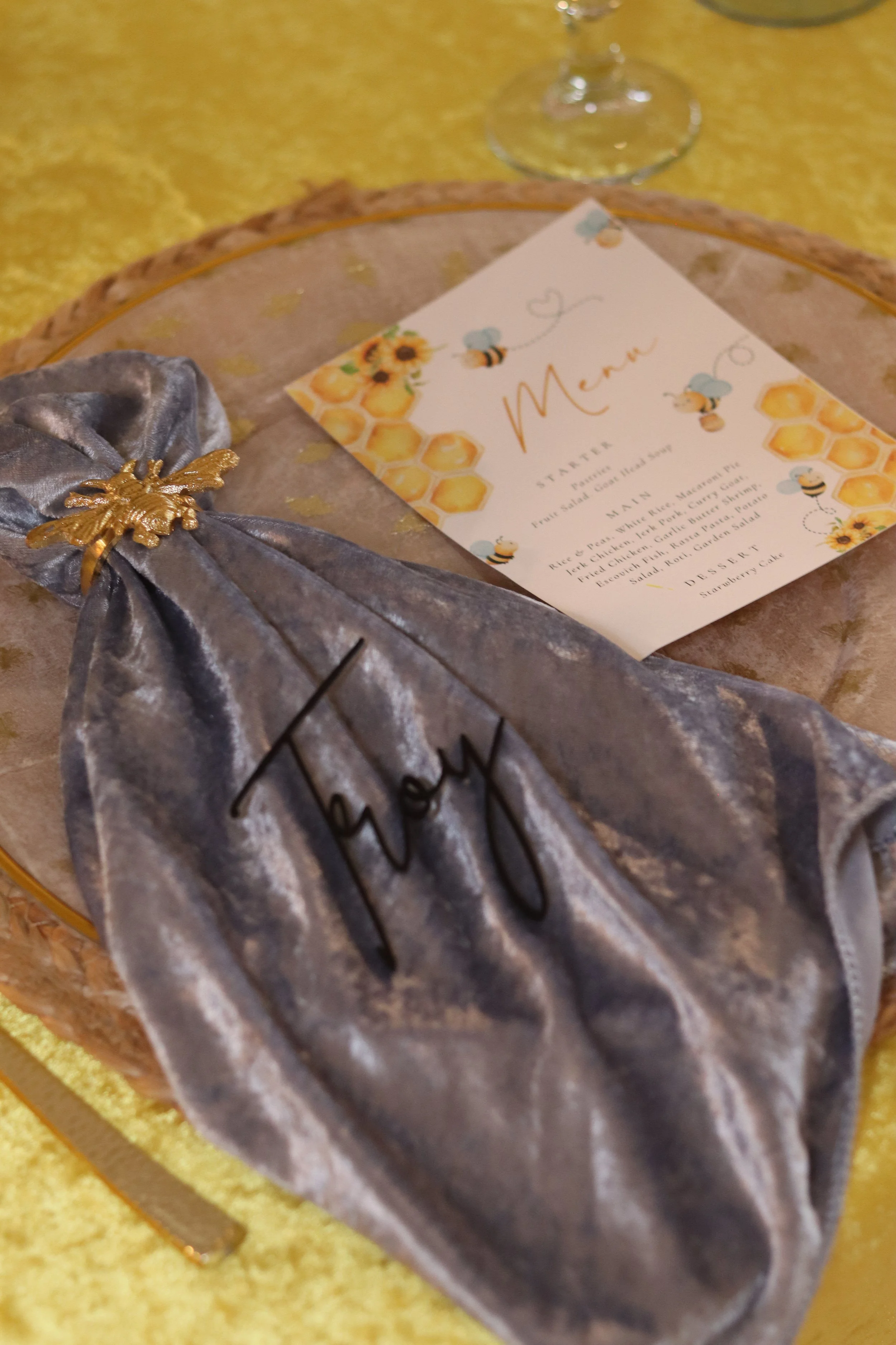 A silver velvet napkin with a gold bee embellishment and the name 'Hoy' embroidered on it, placed on a clear glass charger plate with a honey-themed menu card underneath.