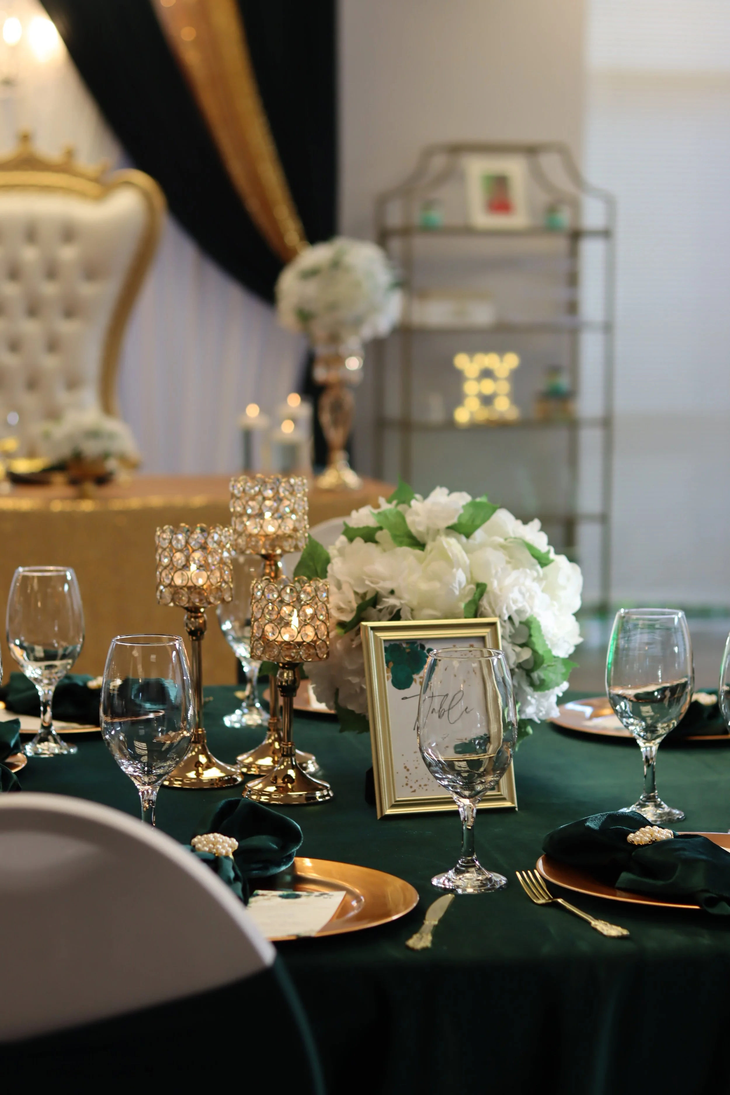 Elegant banquet table with white floral centerpiece, wine glasses, gold charger plates, dark green napkins with pearl napkin rings, and gold candle holders with crystals, set for a formal event in a decorated room.