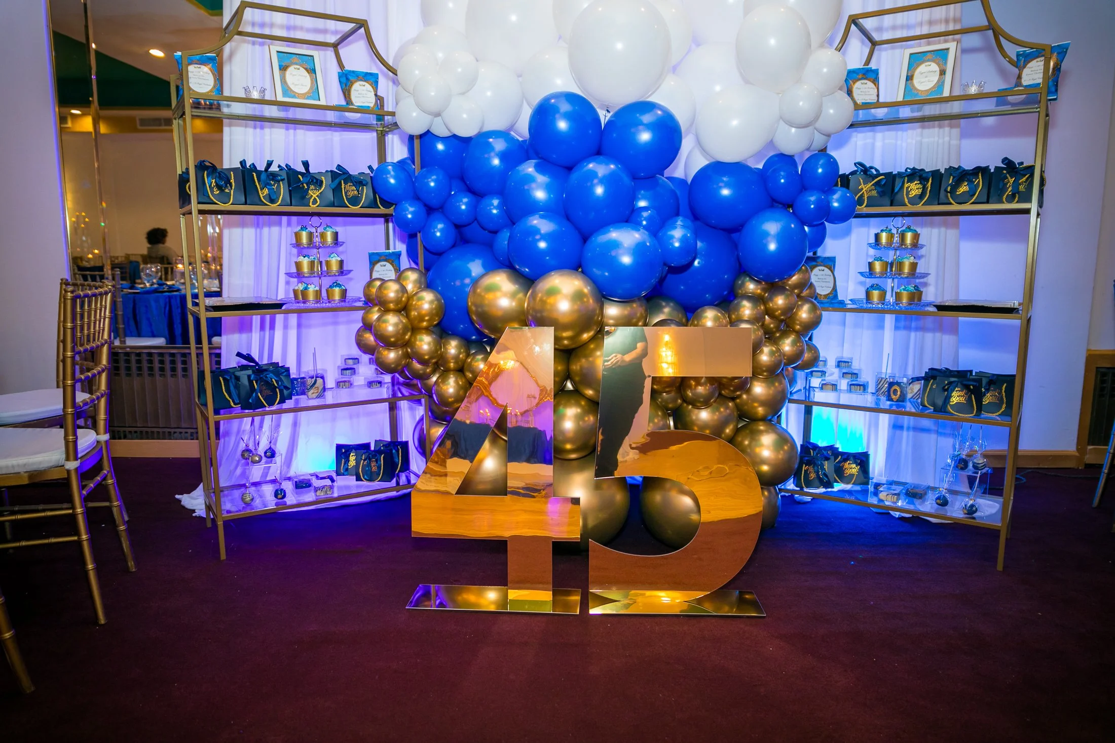 Decorative birthday party display with blue, white, and gold balloons, a large gold '45' sign, and gift bags on shelves.