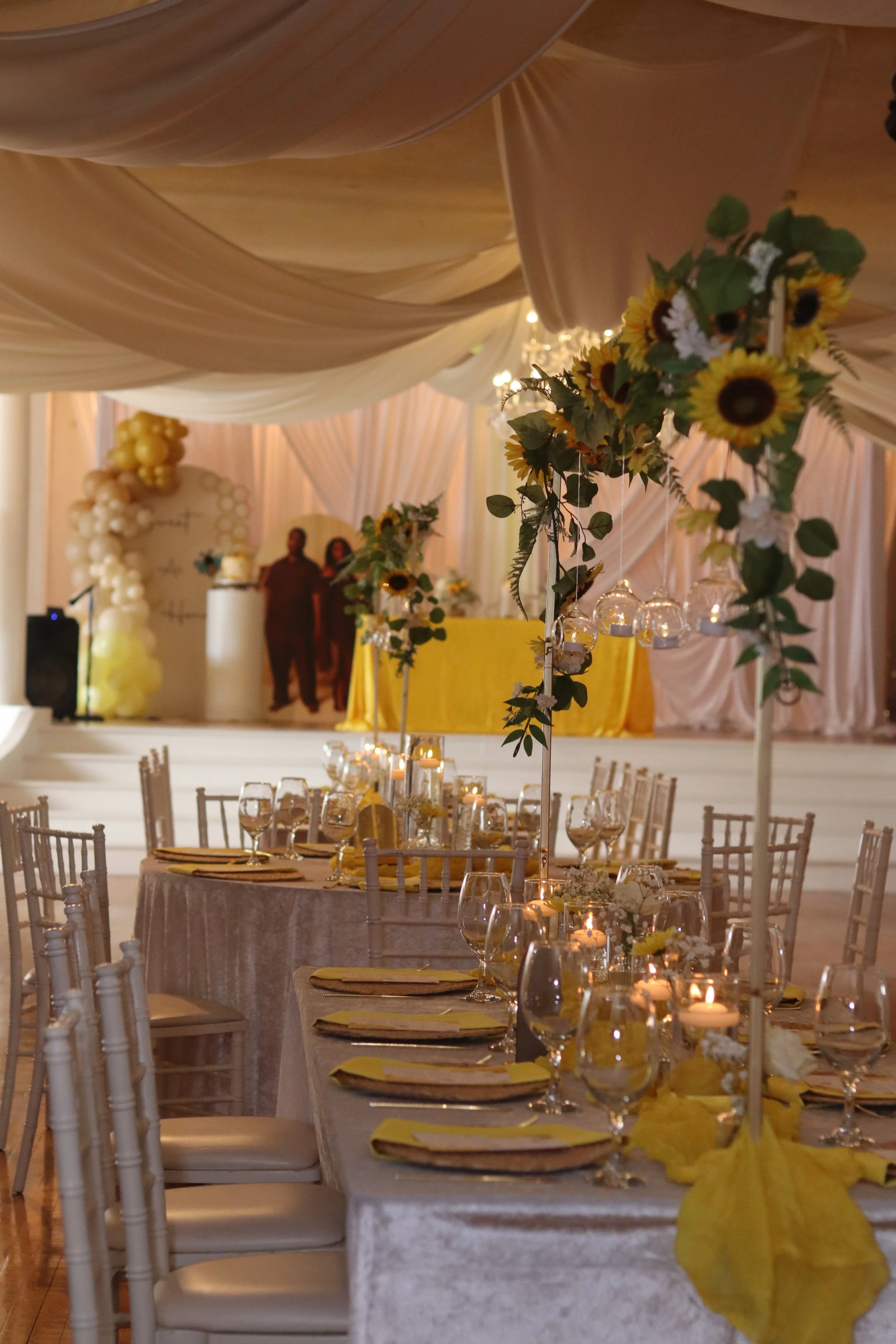 Elegant event space with tables decorated with sunflowers and candles, draped ceiling, and a stage with a yellow tablecloth in the background.