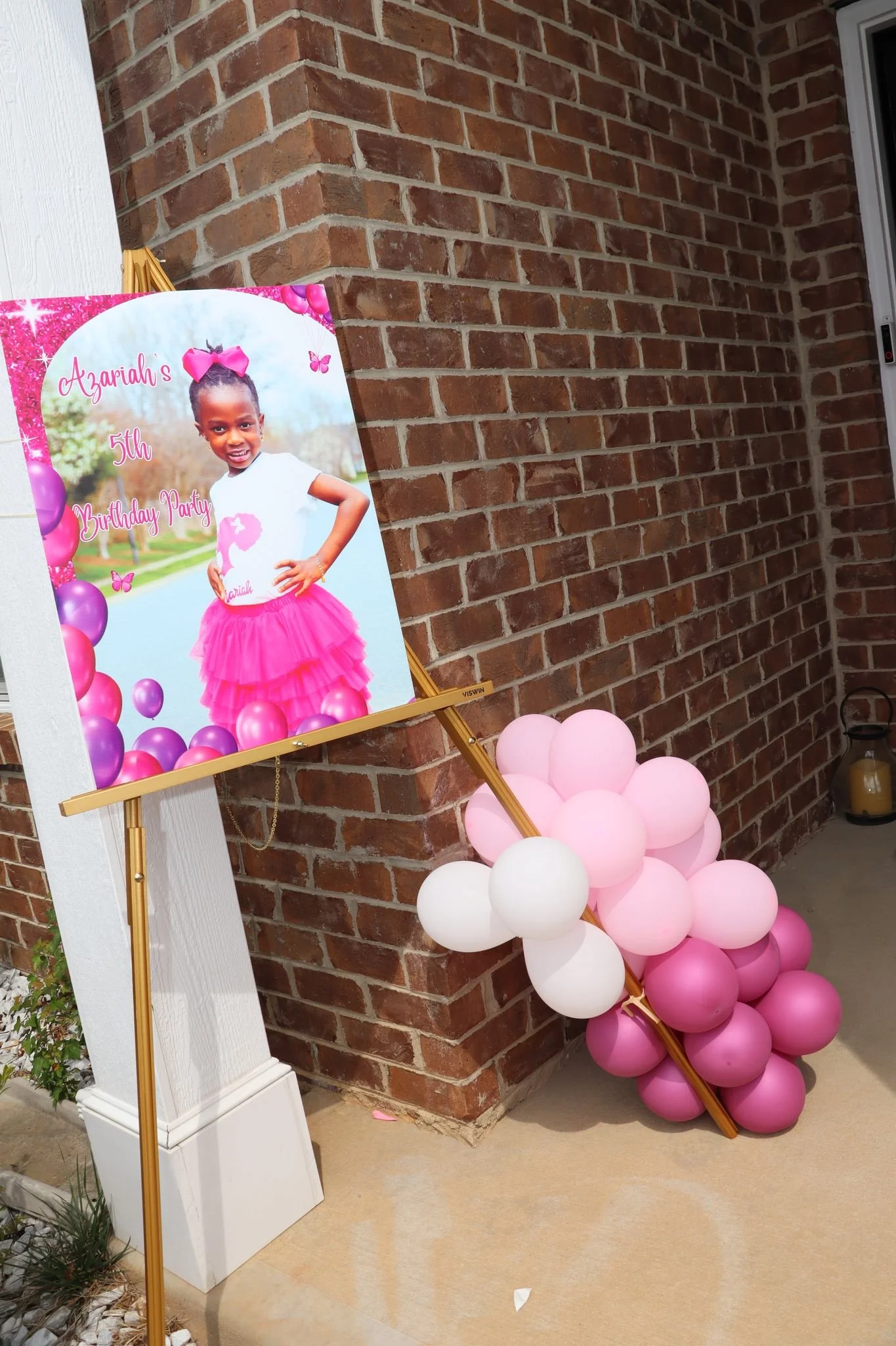 A decorated outdoor area with a birthday poster of a young girl in a pink tutu, pink bow, and white top, celebrating her 5th birthday, with pink and white balloons arranged on the ground.