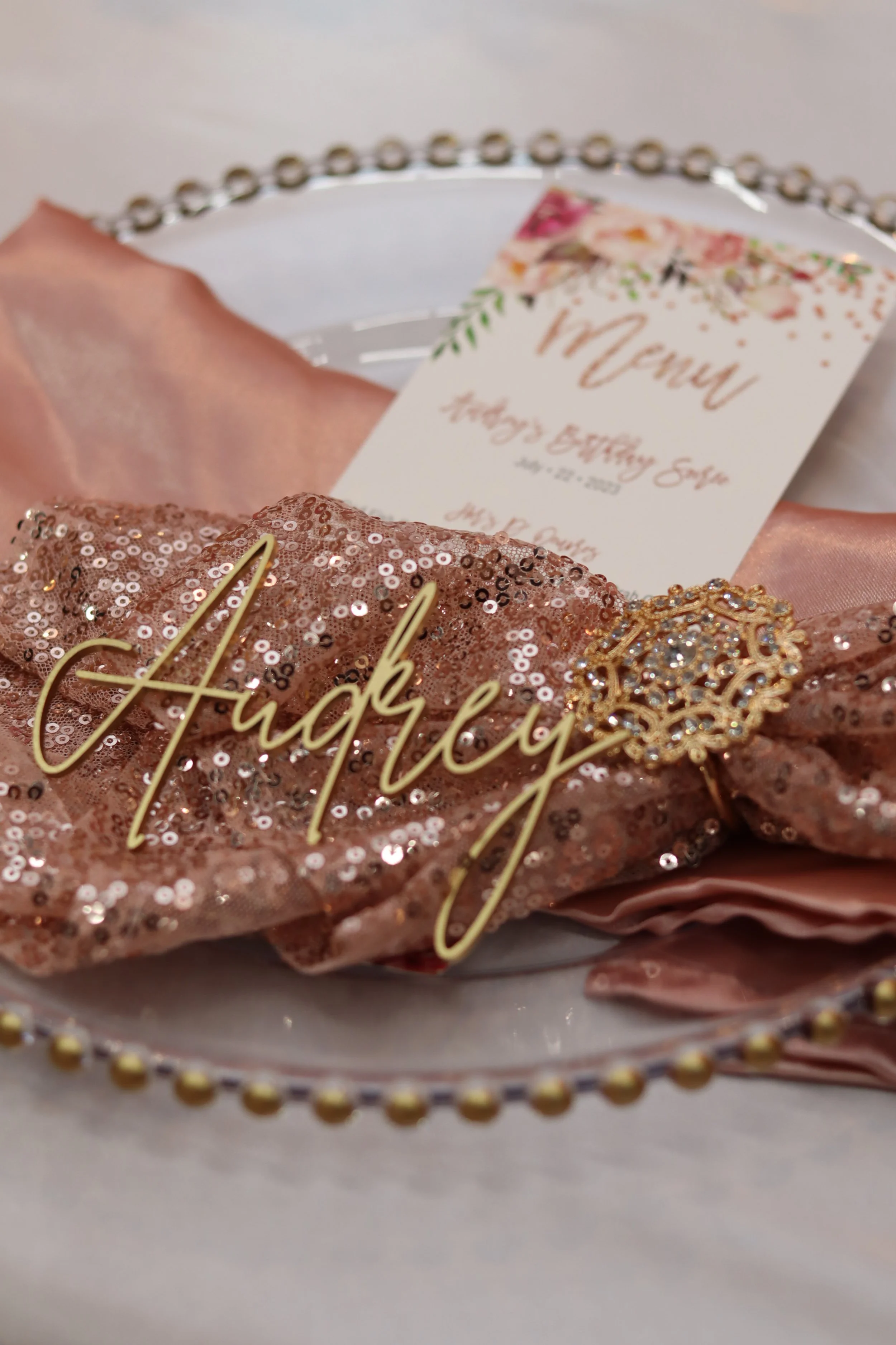 A decorative plate with a pink sequin fabric and a gold necklace spelling 'Aubrey,' along with a gold decorative brooch. In the background, there is a pink satin cloth and a menu card for Aubrey's birthday party.