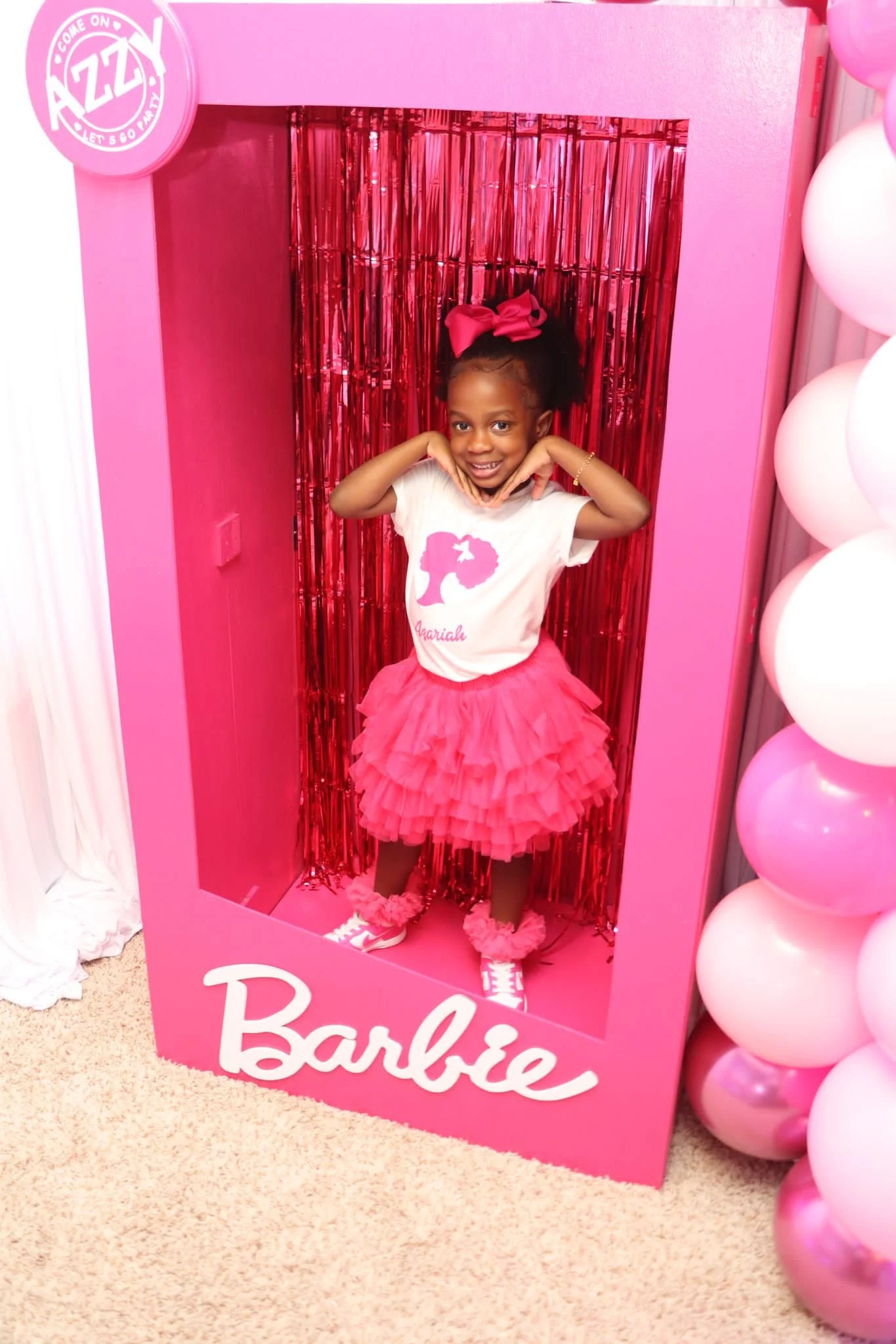 A young girl smiling inside a pink Barbie-themed photo booth with red fringe backdrop, wearing a white T-shirt with a pink silhouette and the name 'Ariella,' paired with a fluffy pink tutu and pink sneakers.