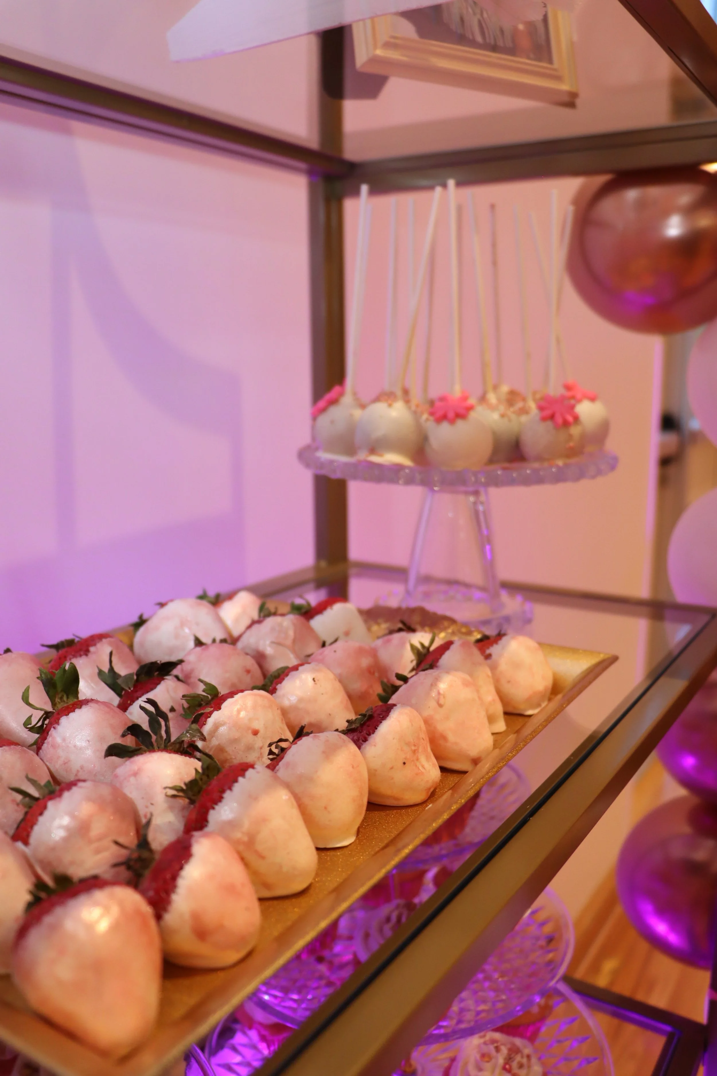 A tray of dipped strawberries and a display of cake pops decorated with pink flowers on a glass tier.