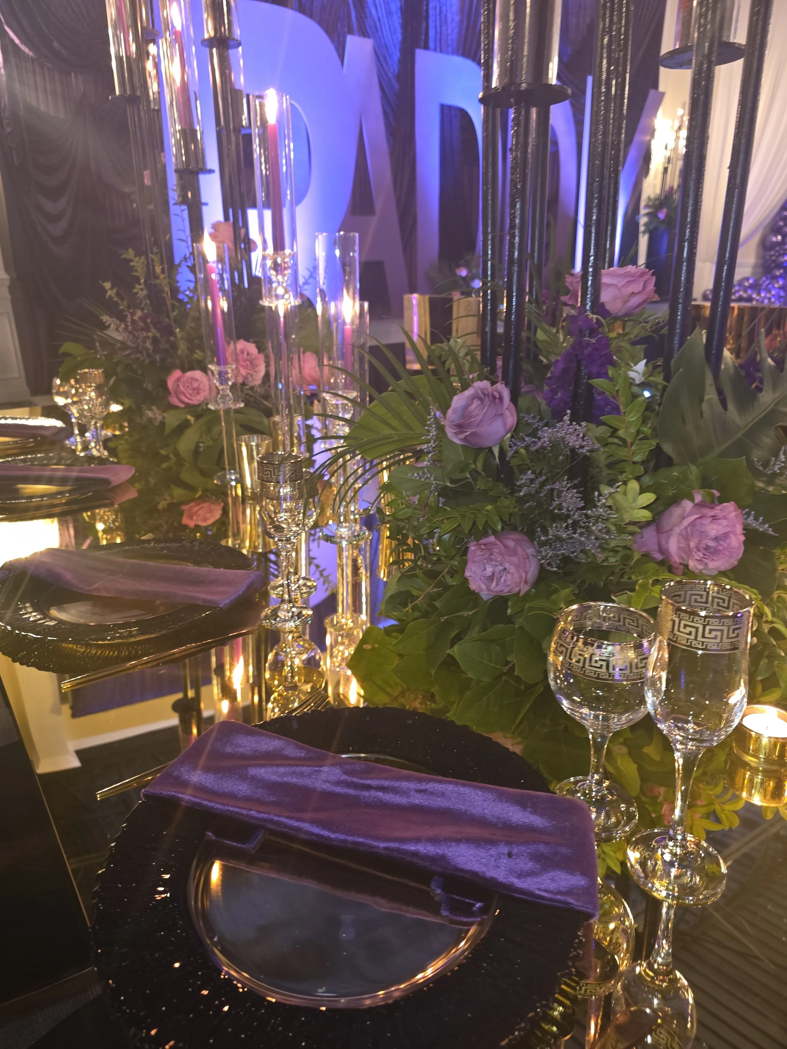 Elegant banquet table setting with purple roses, green foliage, tall candles, glassware, and purple napkins, with a large illuminated sign in the background.