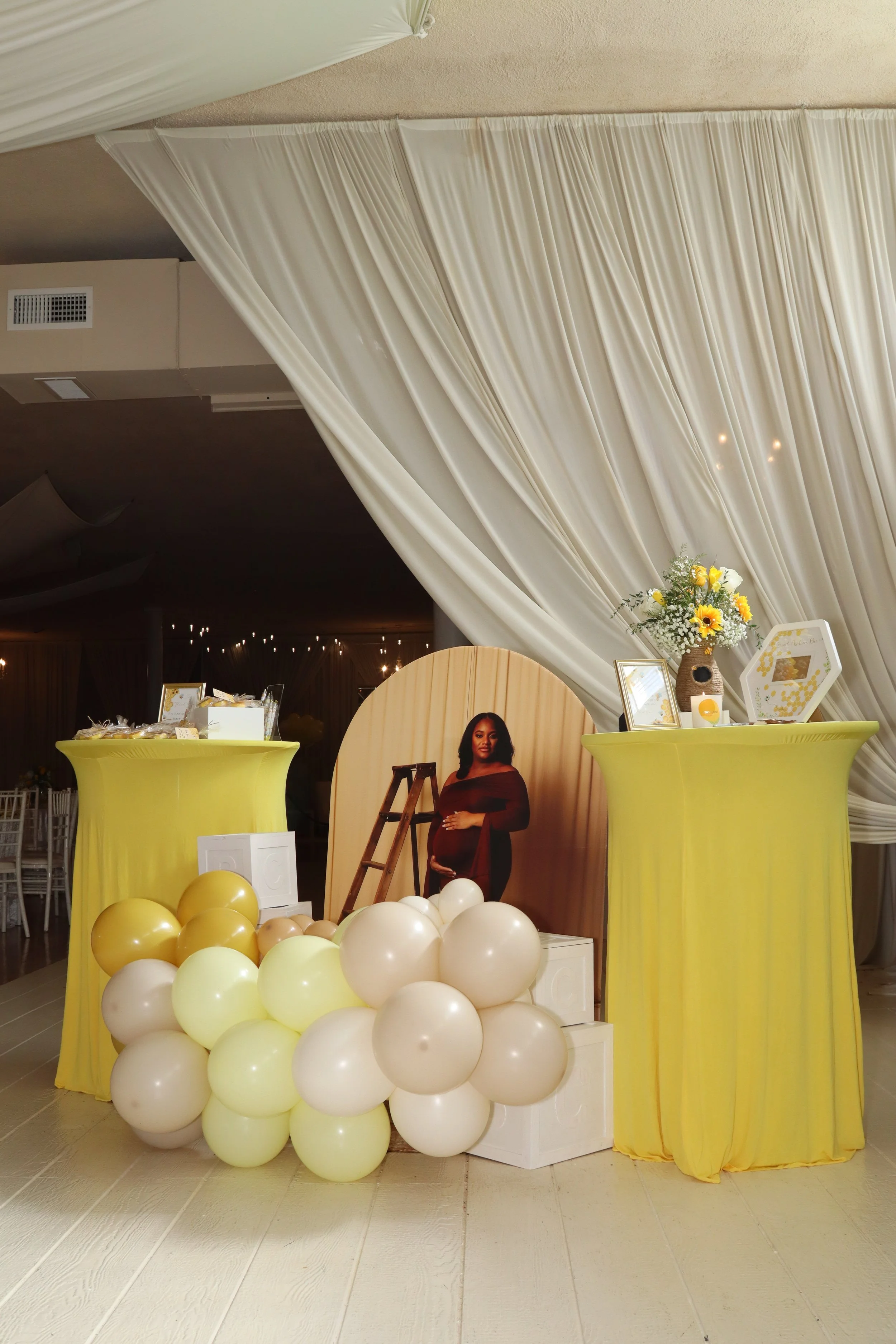 Decorative baby shower display with a standee of a pregnant woman, yellow and white balloons, yellow high tables, a flower arrangement, and framed photos against cream curtains.