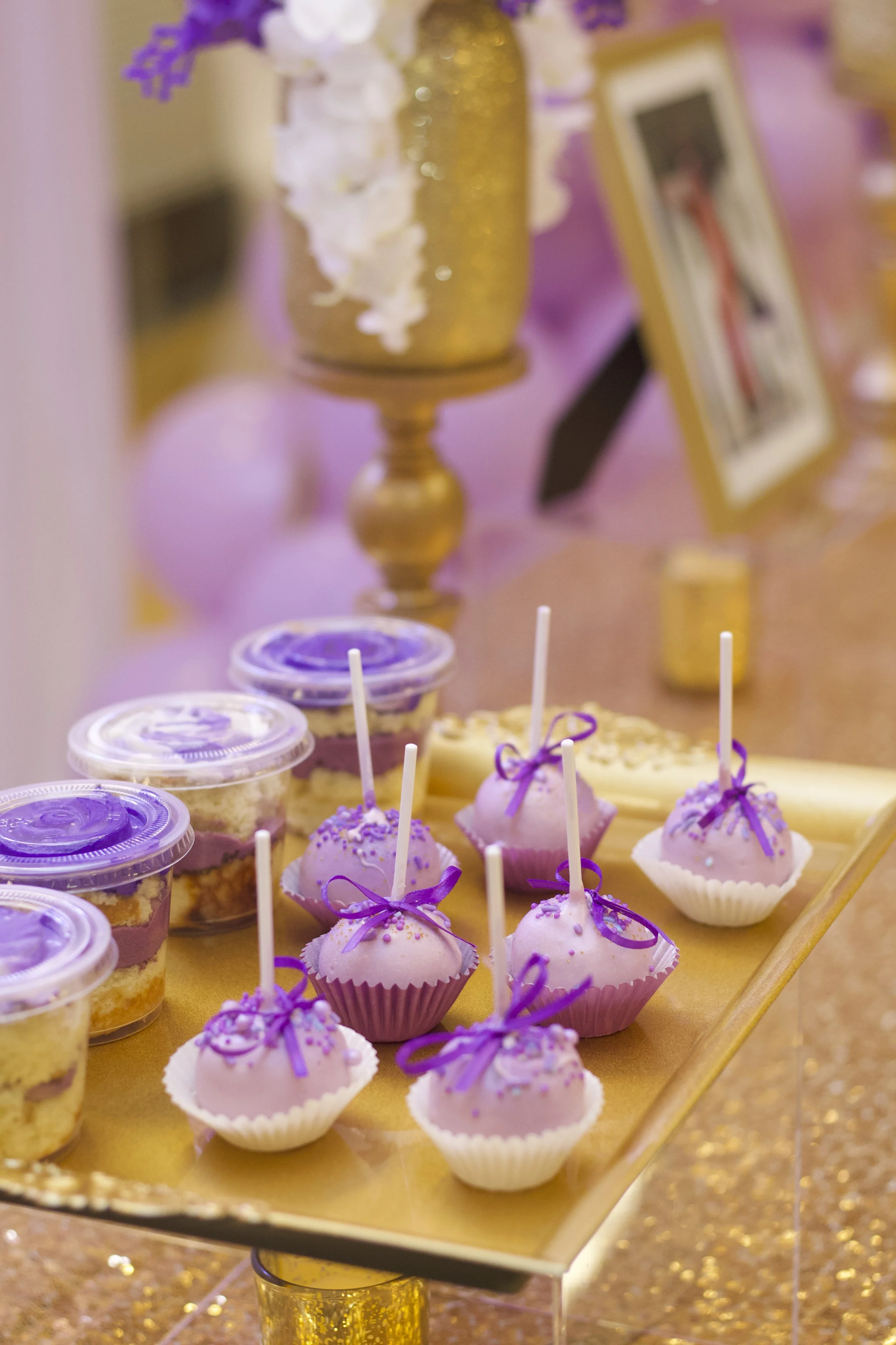 Purple and white decorated cupcakes and dessert cups with purple ribbons on a gold tray at a celebration party.