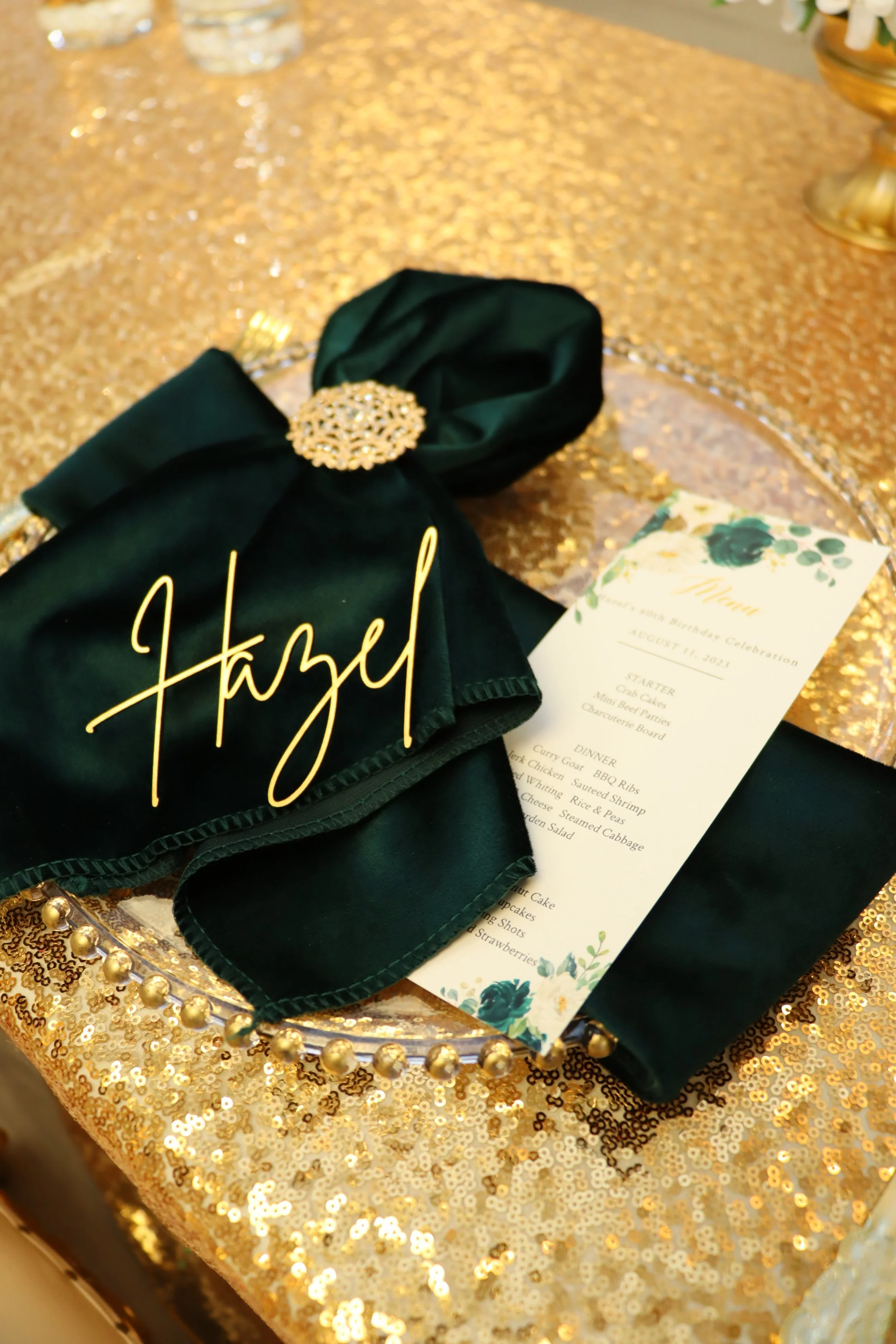 Gold sequin tablecloth with a clear glass charger plate, a green napkin with a gold ring, a menu card, and gold table decorations.