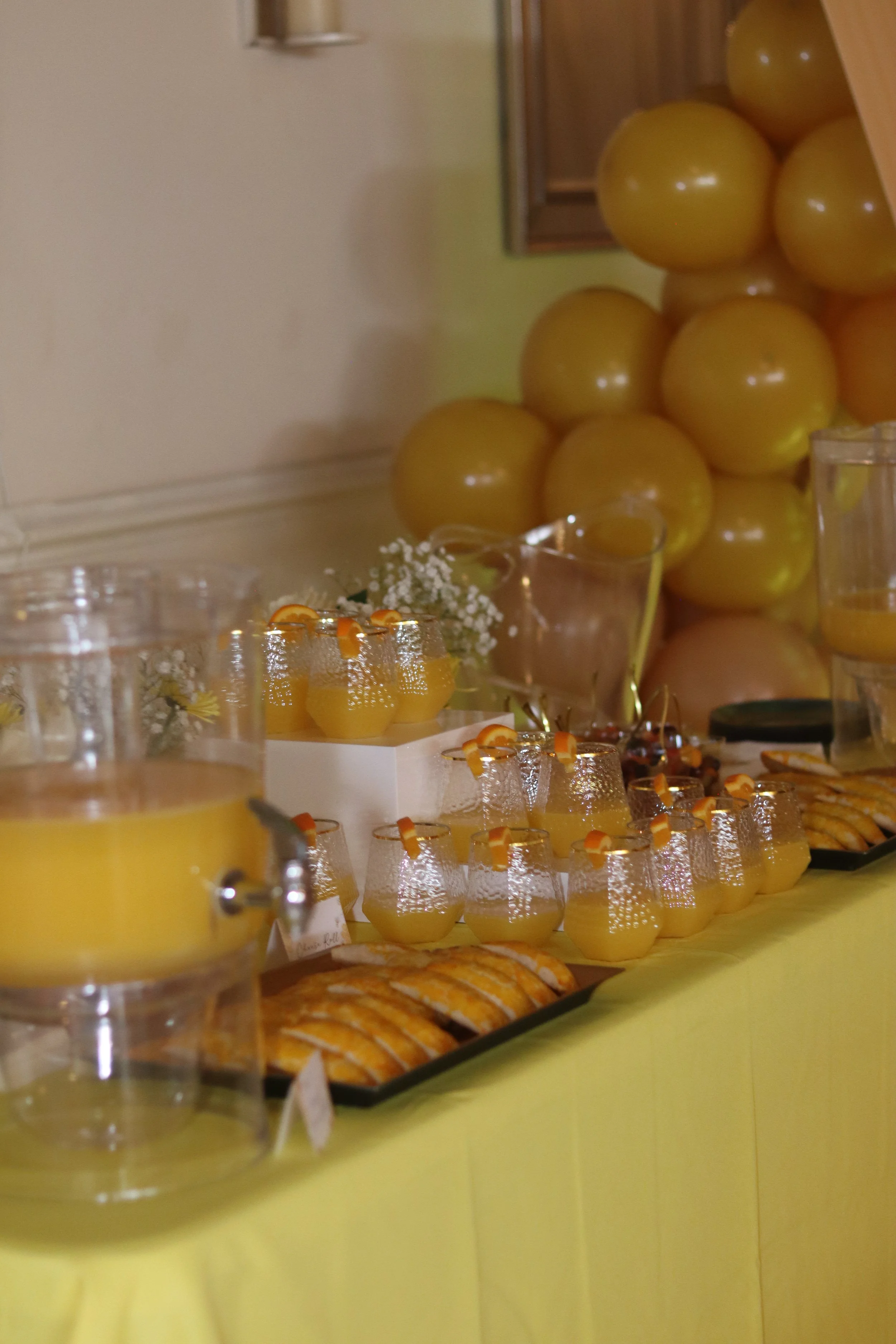 A table with yellow drinks in glasses, candied orange slices, table decorations with foxglove flowers, and a backdrop of large yellow balloons.