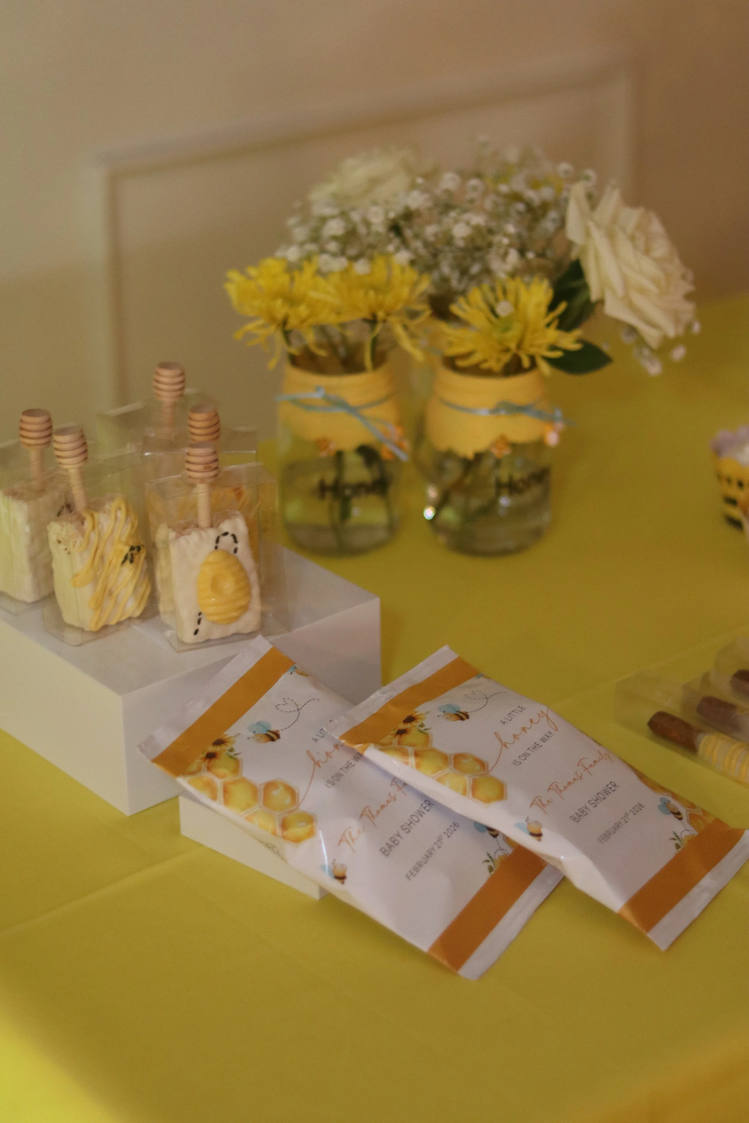 Decorative party favors with honeycomb theme for a baby shower, including honey-themed cookies, floral arrangements in mason jars with yellow and white flowers, and favor bags with honeycombs and bees.
