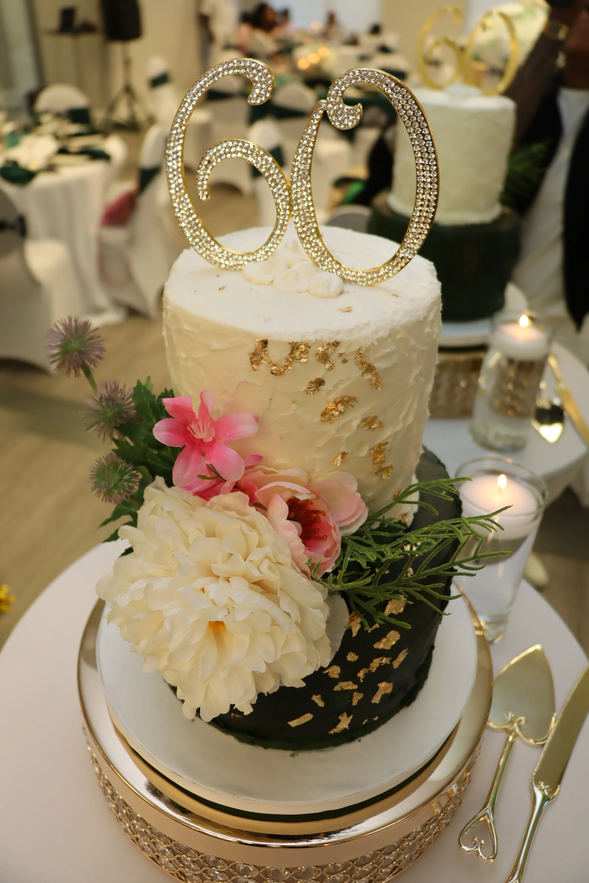 A two-tiered wedding cake decorated with flowers and gold accents, topped with a glittery number 60 cake topper, on a gold and white cake stand surrounded by candles and elegant silverware.