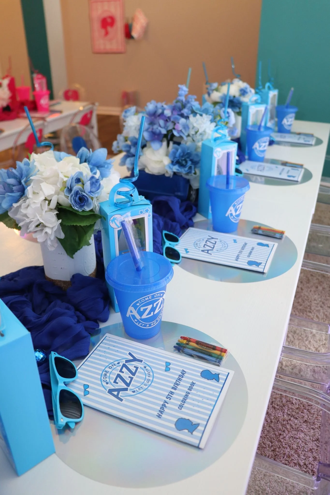 Party table setting for a birthday with blue, white, and purple decorations, including flowers, sunglasses, colorful markers, blue cups with straws, and birthday activity booklets.
