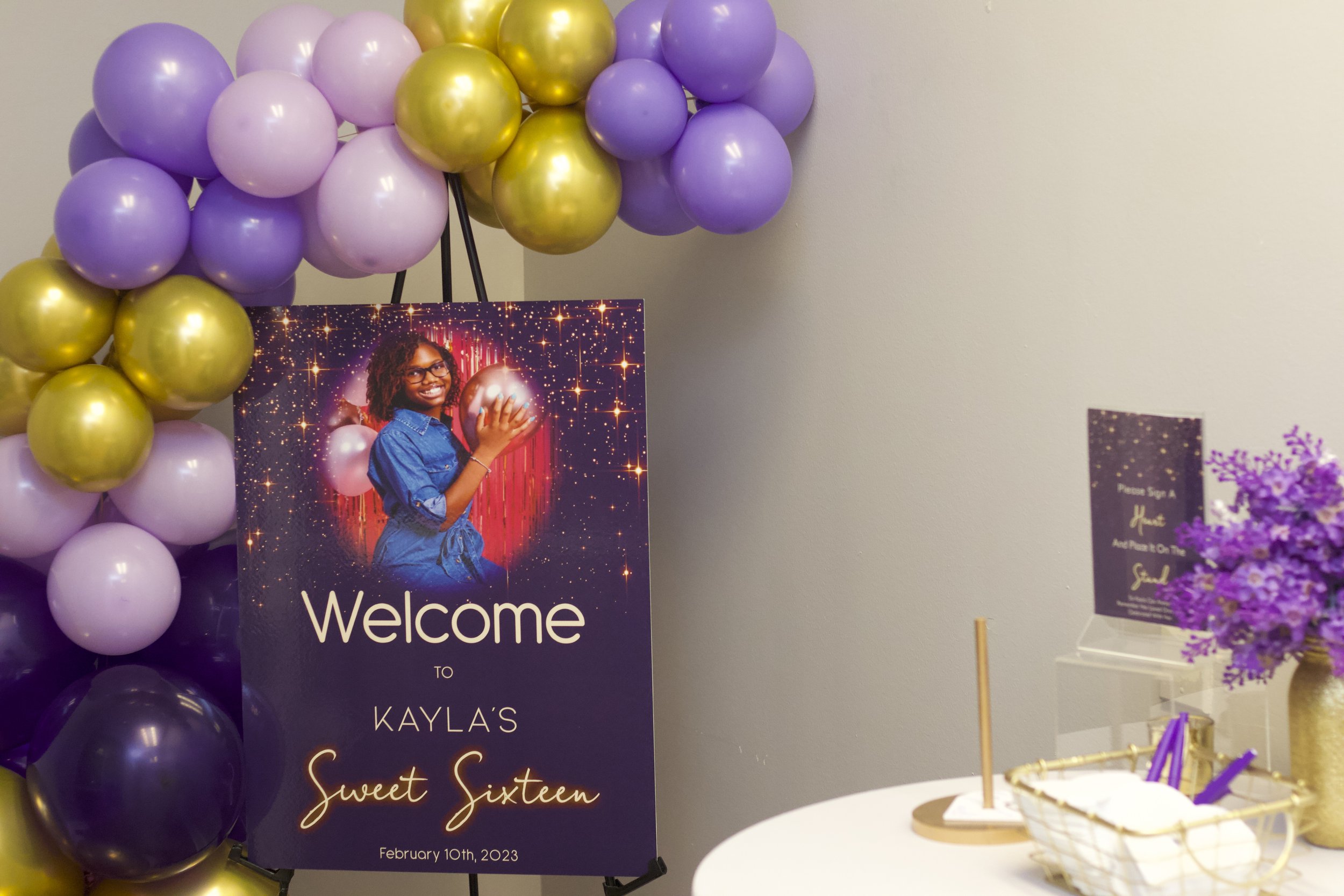 A decorated welcome sign for Kayla's 16th birthday surrounded by purple and gold balloons, with a table on the right holding purple flowers, a sign-in sheet, and purple pens.