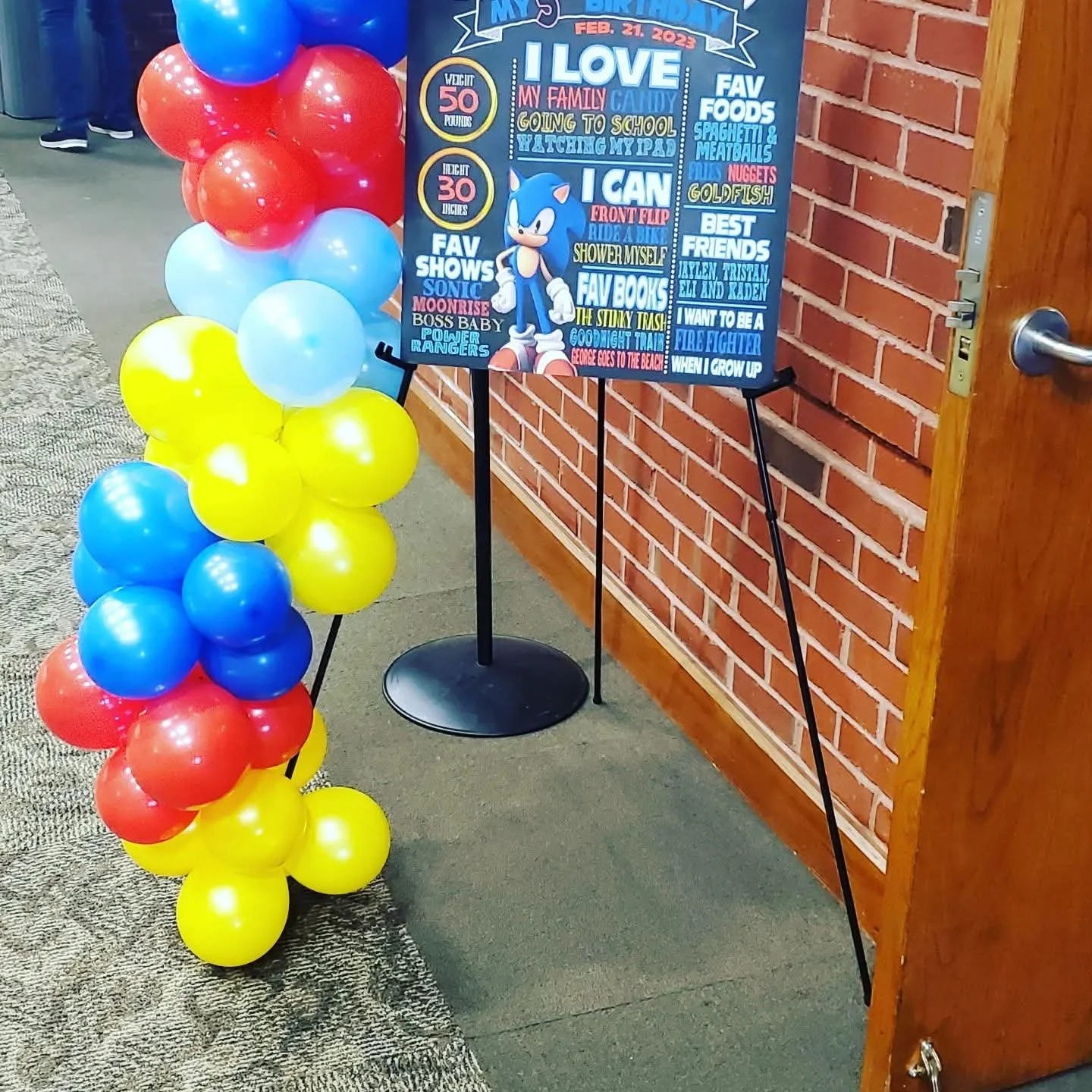 Decorative sign celebrating a child's birthday with colorful balloons attached to it, including red, yellow, blue, and light blue balloons, positioned next to a brick wall and a wooden door.
