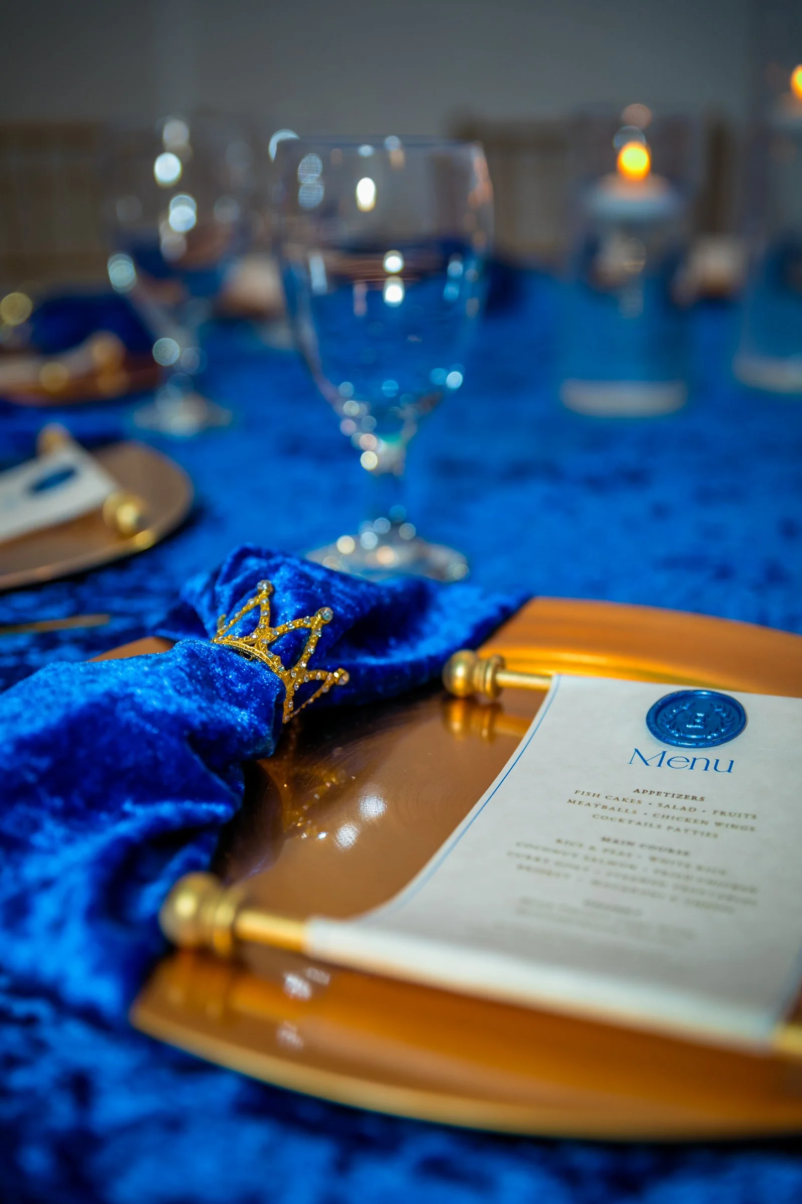 A banquet table decorated with a royal blue velvet tablecloth, gold accents, a menu, a glass of water, and a small gold crown decoration.