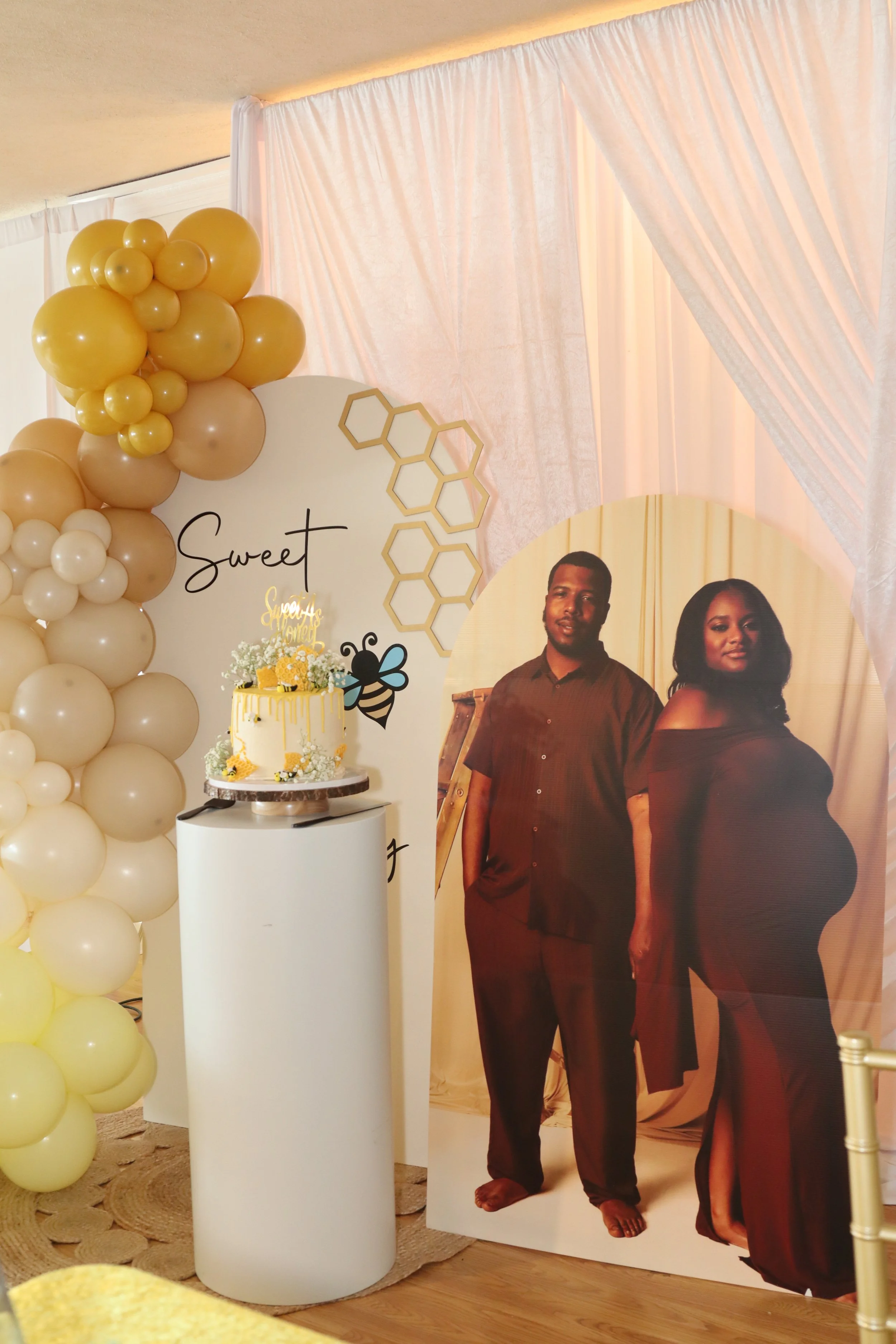 A celebration setup with a blonde man and woman by a pregnant woman in a black off-shoulder gown, a cake on a white pedestal, balloons in cream and gold, and decorative honeycomb and bee accents, with the word "Sweet" on a white backdrop.
