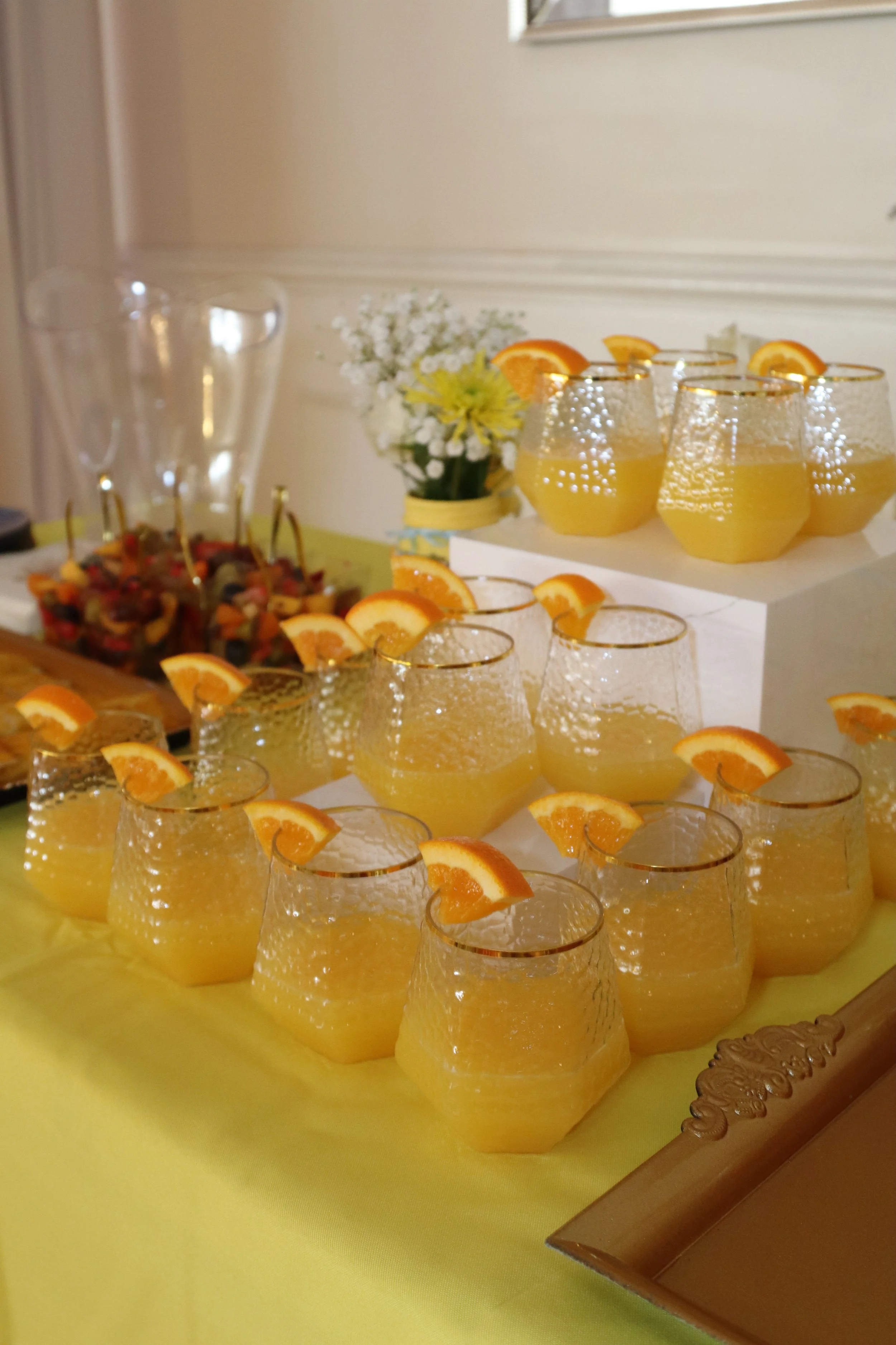 Glassware with orange slices filled with yellow beverage, arranged on a yellow tablecloth at a party or celebration.