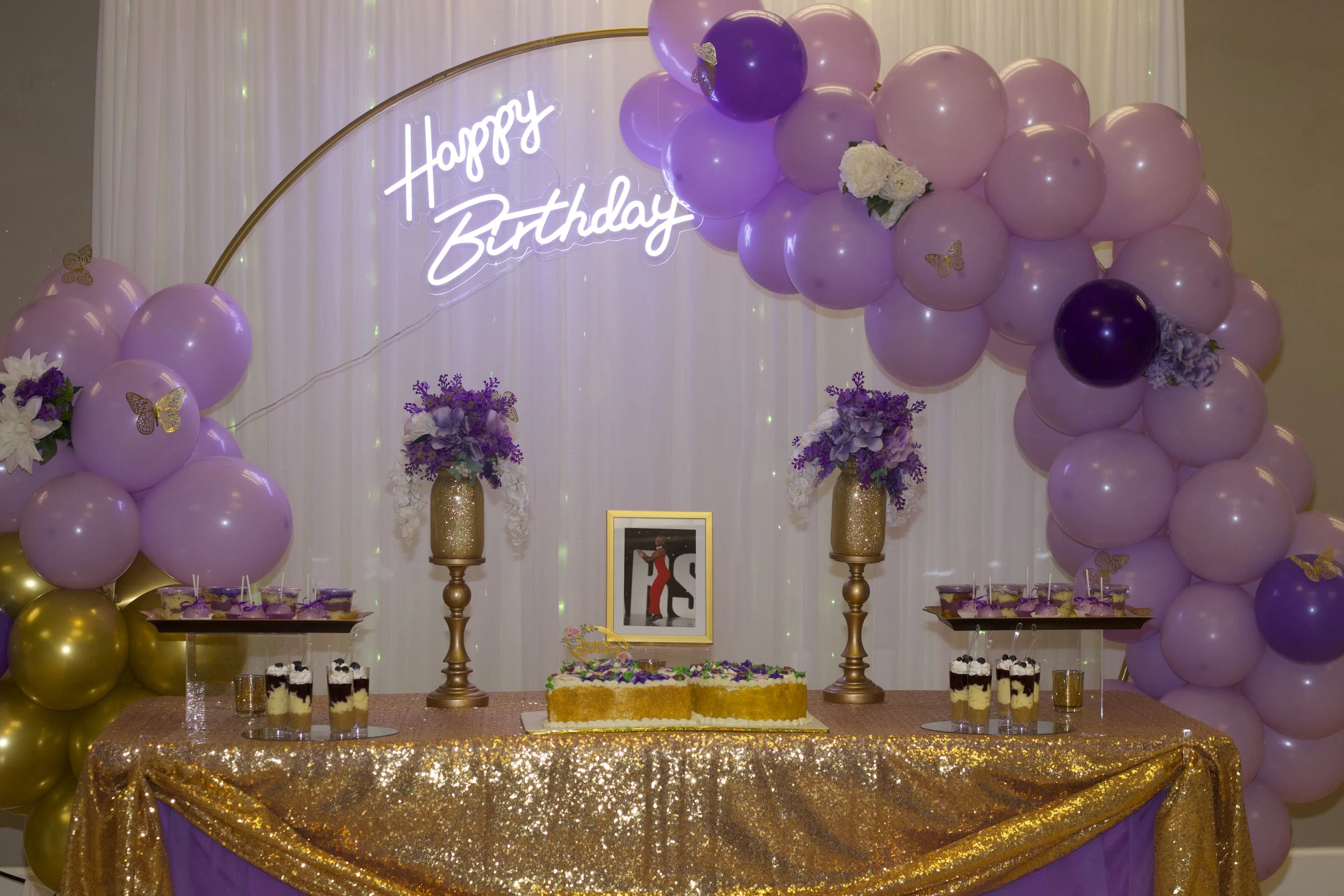 Birthday celebration table decorated with purple and gold balloons, purple flowers, a 'Happy Birthday' neon sign, a cake, and framed photograph.