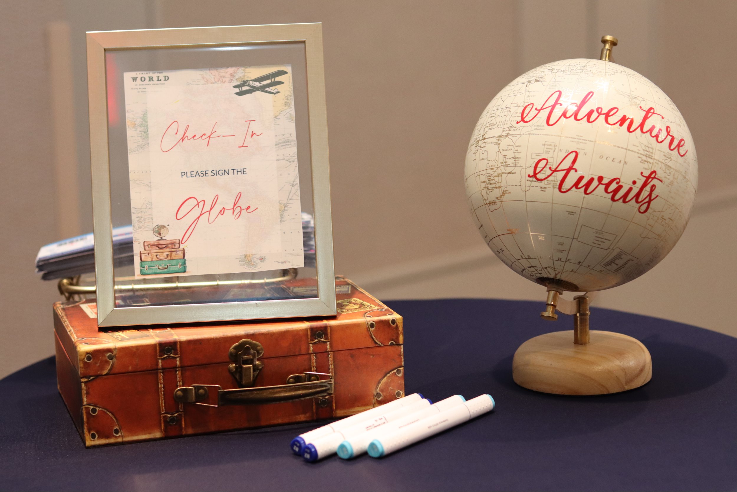 Travel-themed check-in station with a framed sign reading "Check-In" and "Please sign the Globe", vintage suitcase, a globe with the phrase "Adventure Awaits" written on it, and markers on a table.