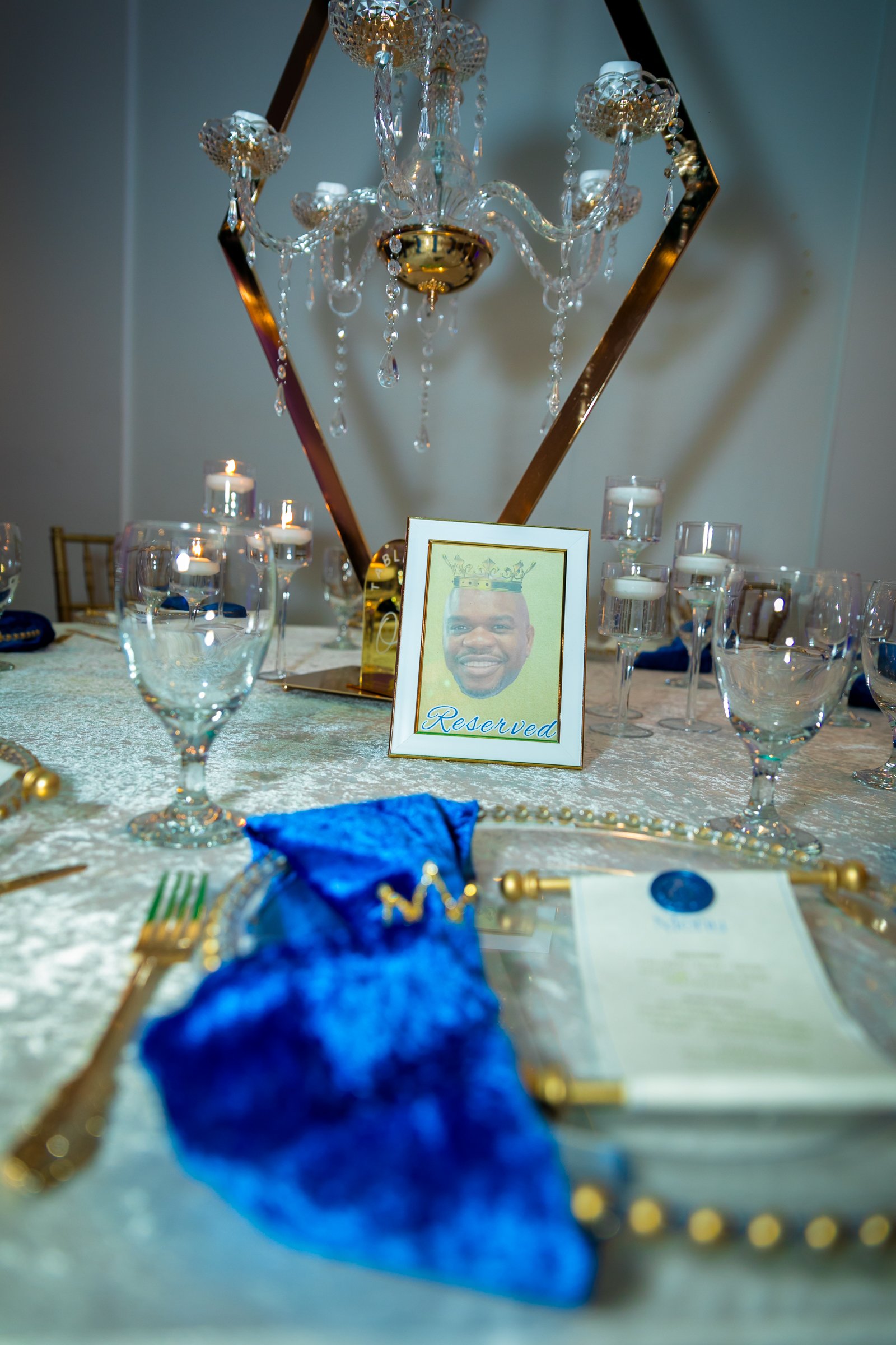 Elegant dining table with crystal glasses, candles, and gold accents, featuring a framed photo of a man with a crown and a 'Reserved' sign. A blue napkin with a gold letter is in the foreground.