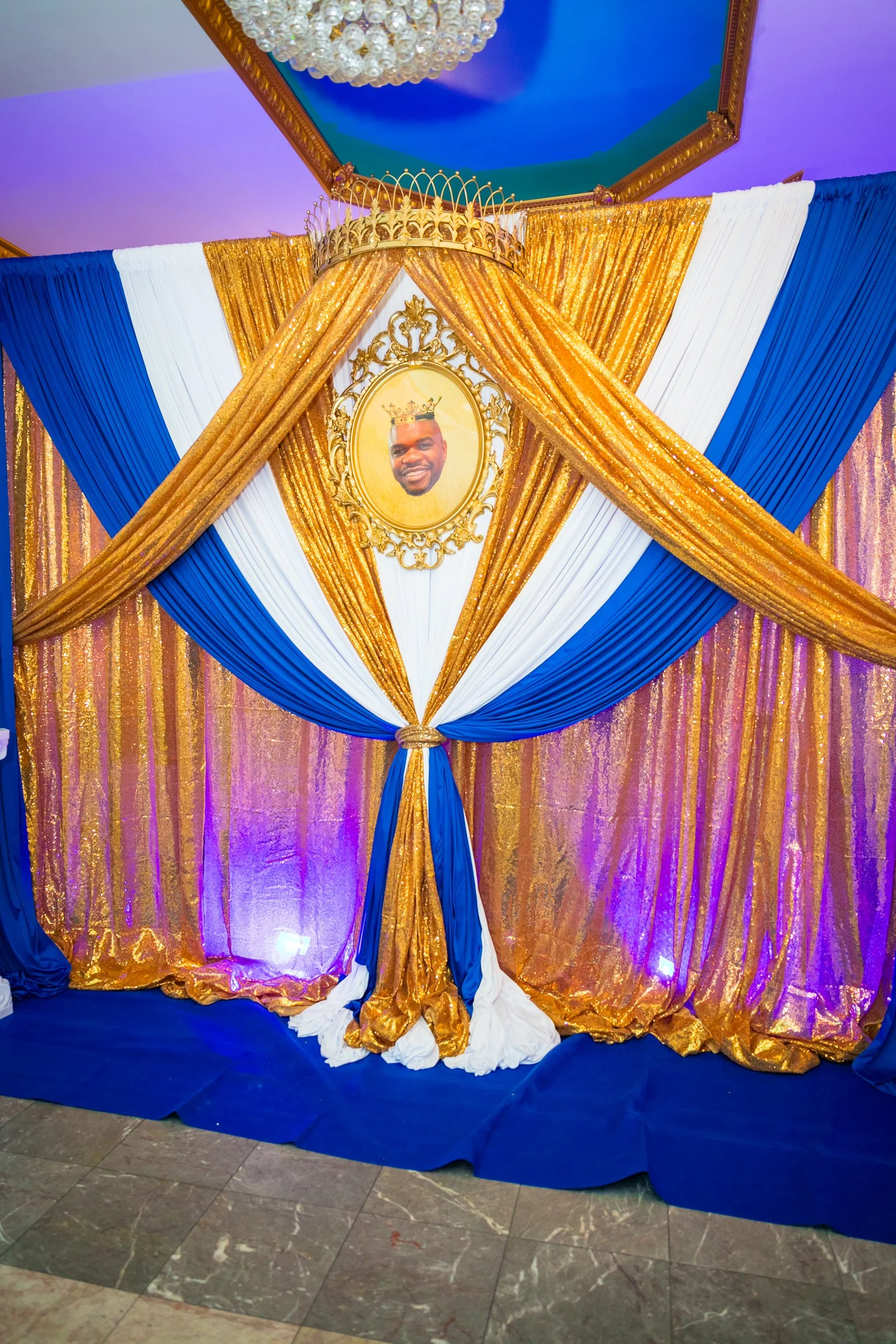 Decorative backdrop with gold, white, and blue drapes, topped with a crown, and a framed photo of a smiling man wearing a crown at a celebration or event.
