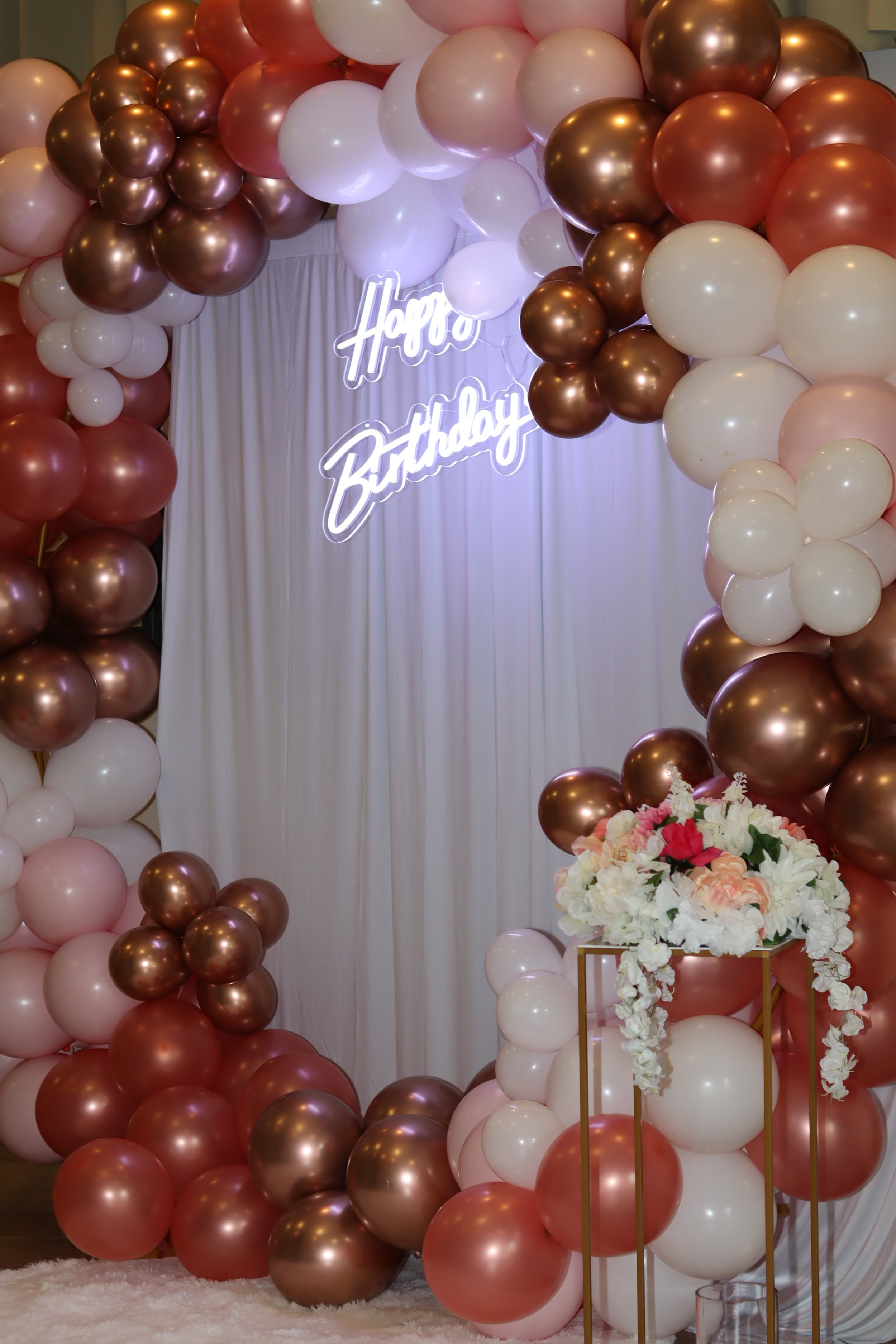 Decorated birthday celebration with balloon arch in pink, white, and metallic rose gold balloons surrounding a white backdrop with a 'Happy Birthday' neon sign and a floral arrangement on a gold stand.