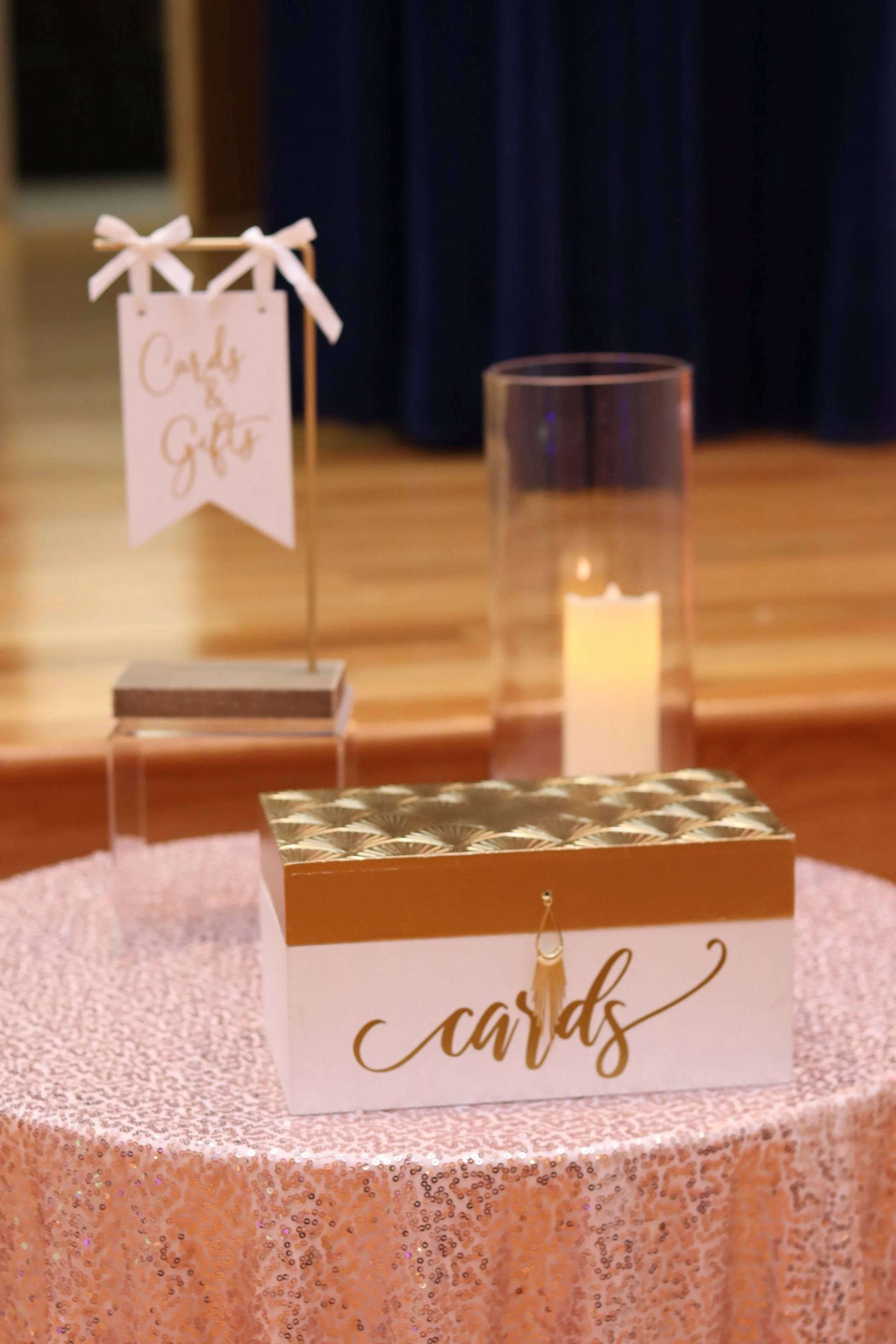 Decorative wedding card box on a pink sequin tablecloth, with a sign that reads 'Cards & Gifts,' a glass candle holder with a lit candle, and a small stand with a 'Cards & Gifts' sign in the background.
