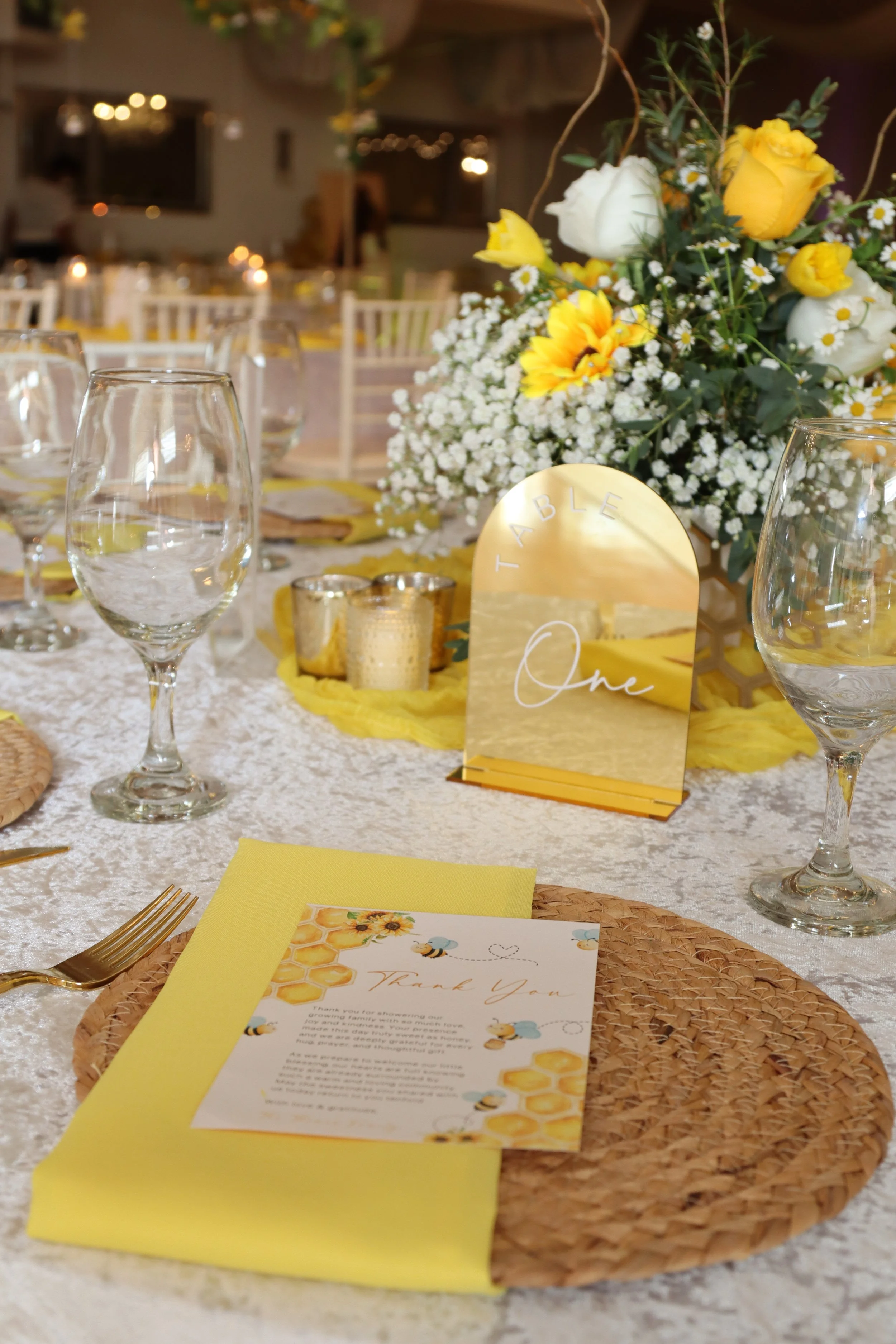 A table setting at a wedding reception with a centerpiece of white and yellow flowers, a mirror with the word 'TABLE' and number 'One', gold candles, clear wine glasses, a woven place mat, yellow napkin, and a thank you card with bee and honeycomb il