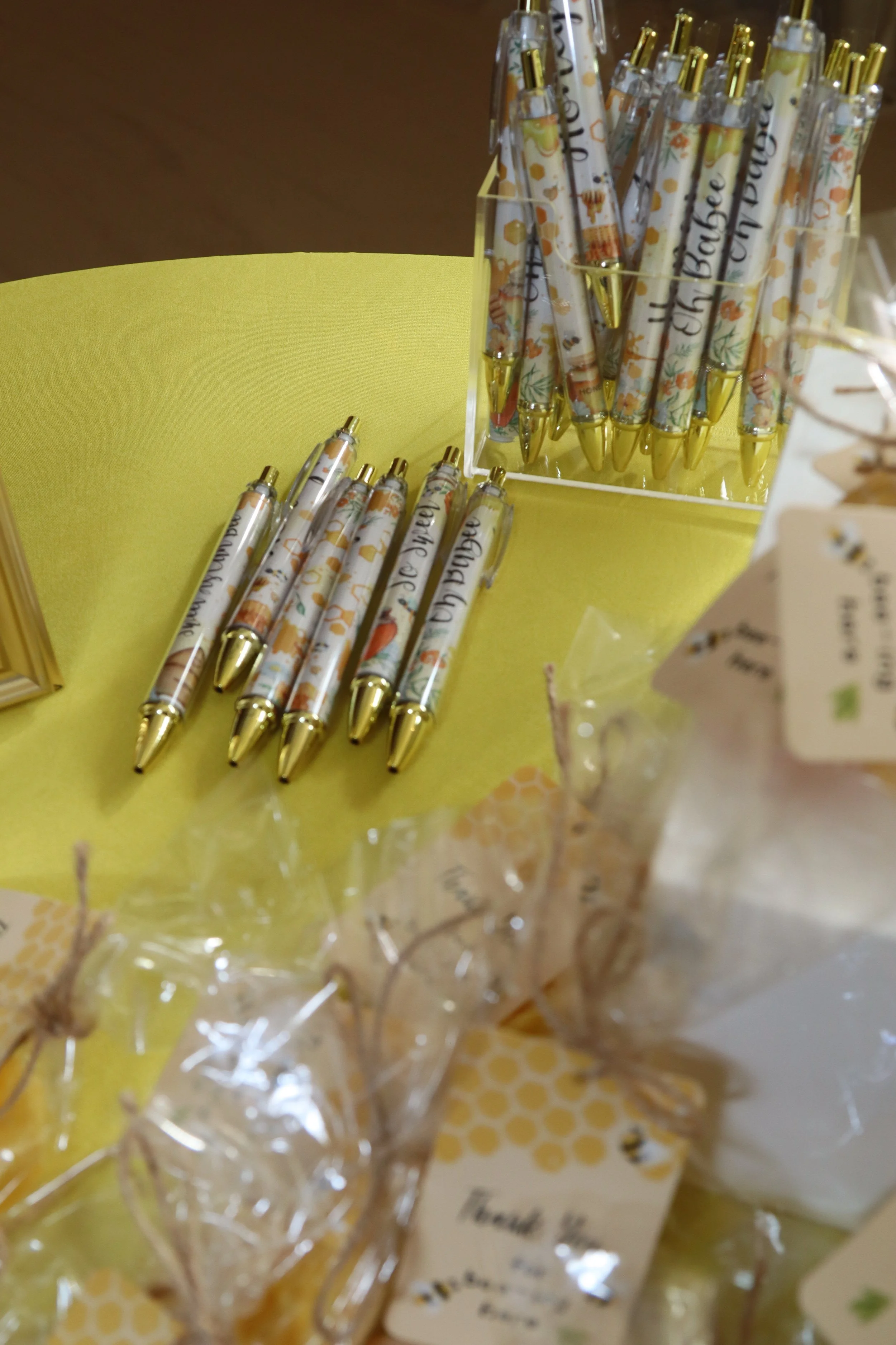 Colorful pens with gold tips and decorative exteriors are displayed on a yellow surface, with a clear container holding more pens in the background.