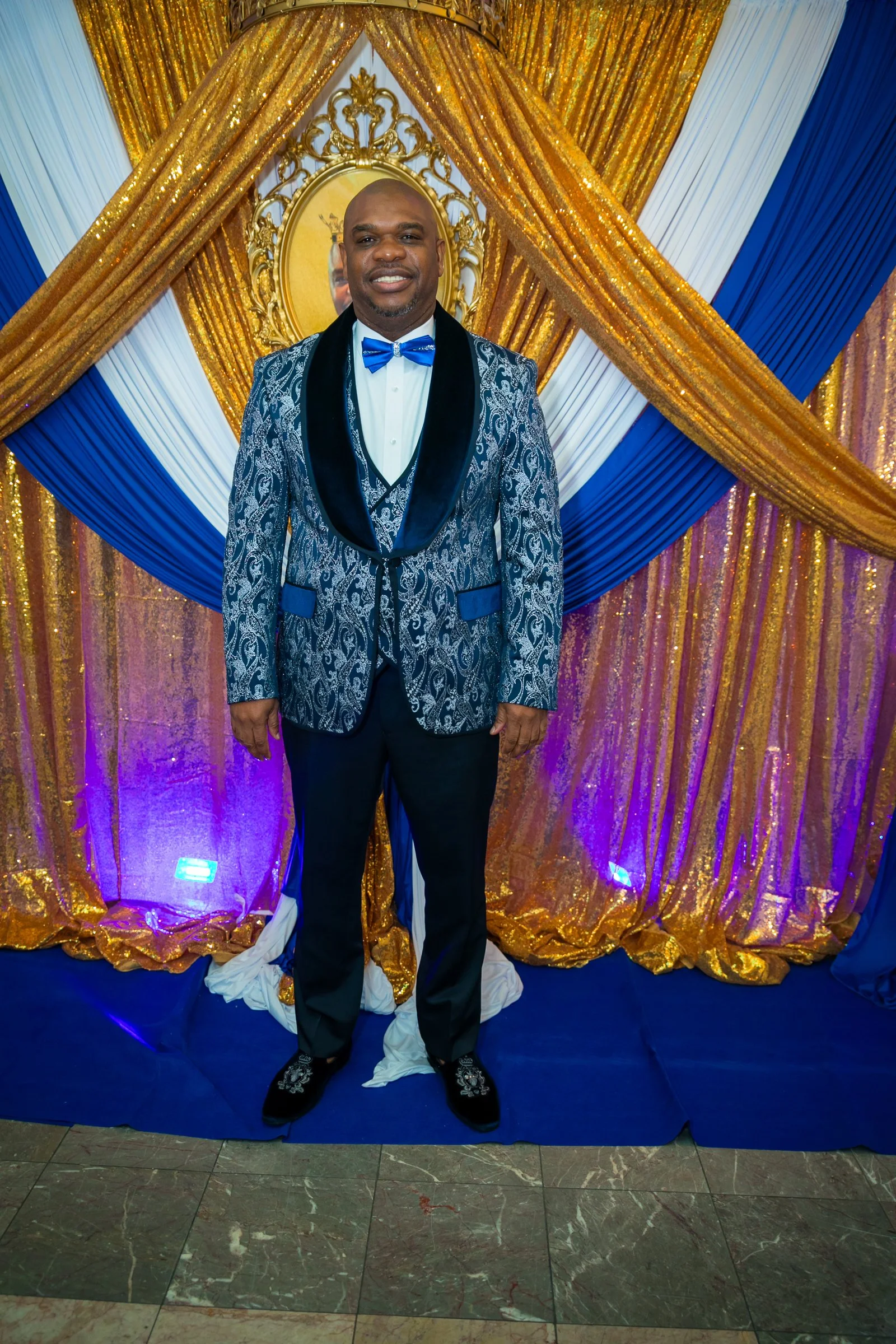 A man in a blue patterned tuxedo with black lapels, white shirt, and blue bow tie standing in front of a gold, white, and blue draped backdrop with a circular gold-framed mirror.