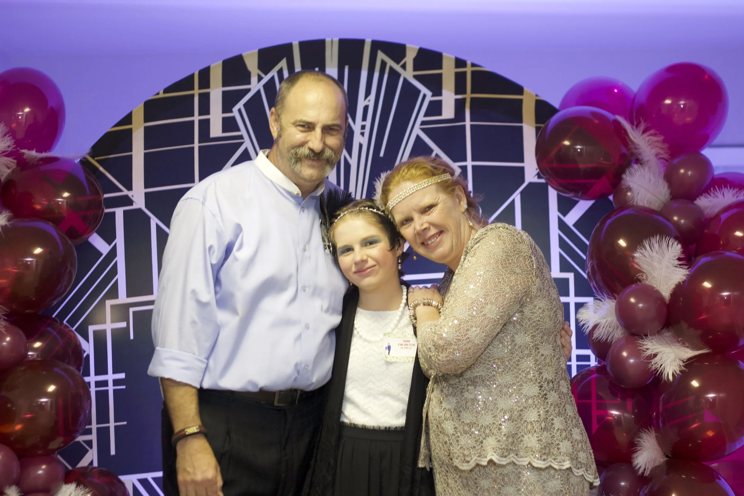 A group of four people, including a man with a beard and a mustache, a woman with a headband, and a young girl, standing together at a social event or celebration, with pink balloons and an abstract black and white backdrop in the background.