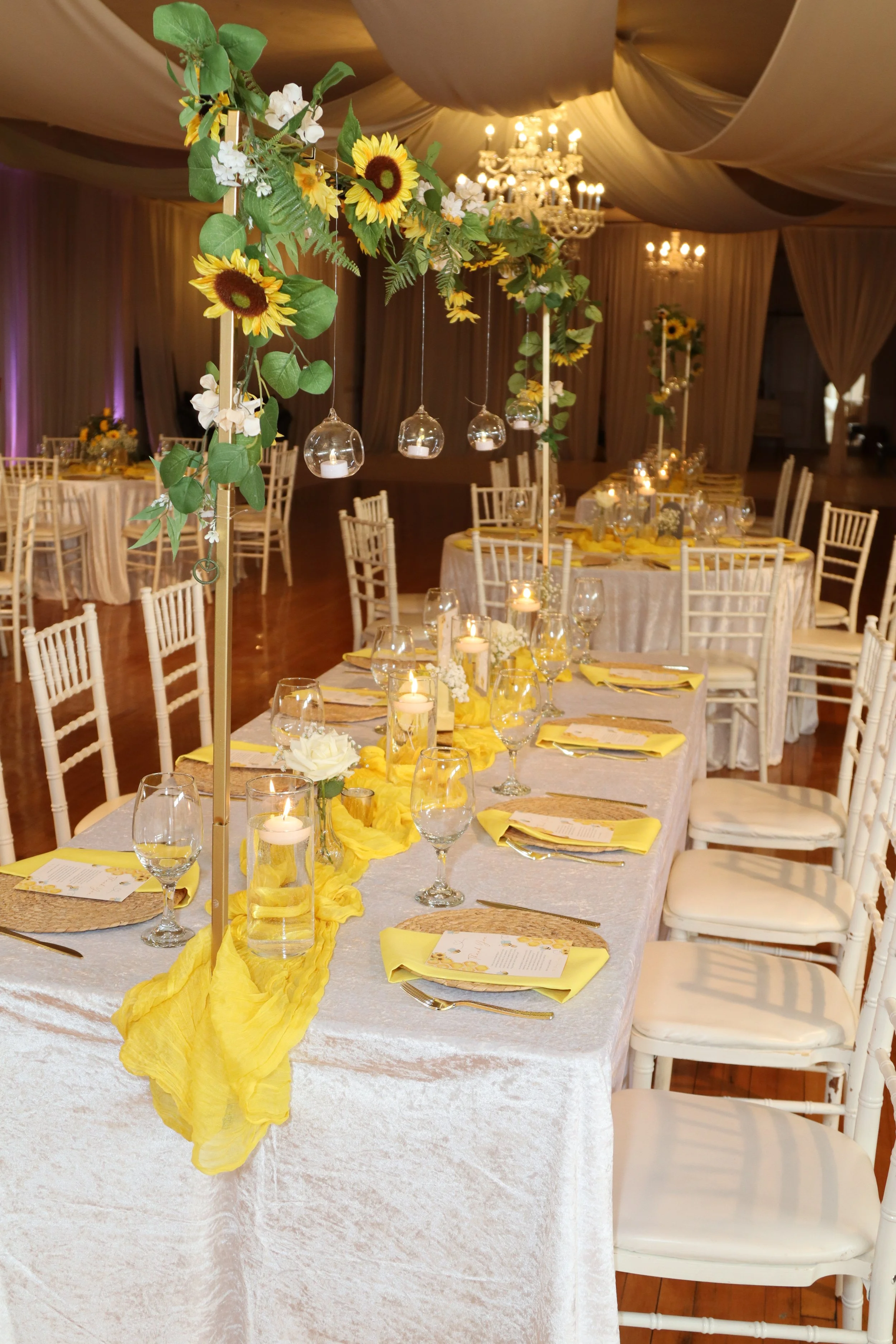 Elegant banquet table decorated with tall sunflower and greenery centerpieces, candles, and yellow accents, set in a decorated ballroom with chandeliers.