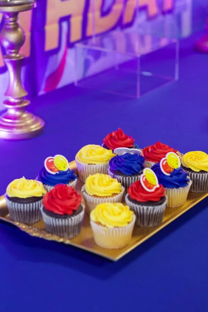 Colorful cupcakes with yellow, red, and blue frosting and decorative toppers on a gold tray, set on a blue table.