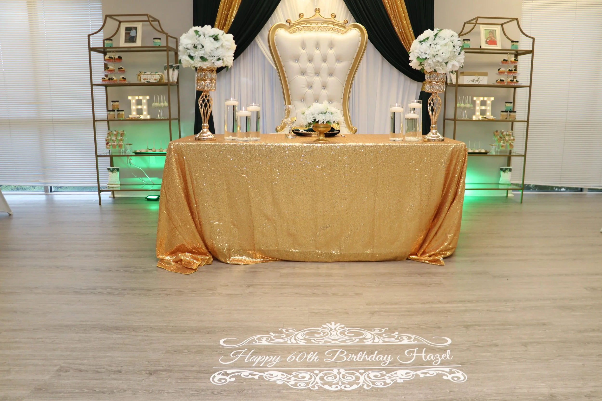 Elegant birthday celebration setup with a gold glitter tablecloth, a large white ornate chair, floral centerpieces, candles, and decorative shelves with framed photos and gifts. The floor has a custom message: 'Happy 60th Birthday Hazel'.