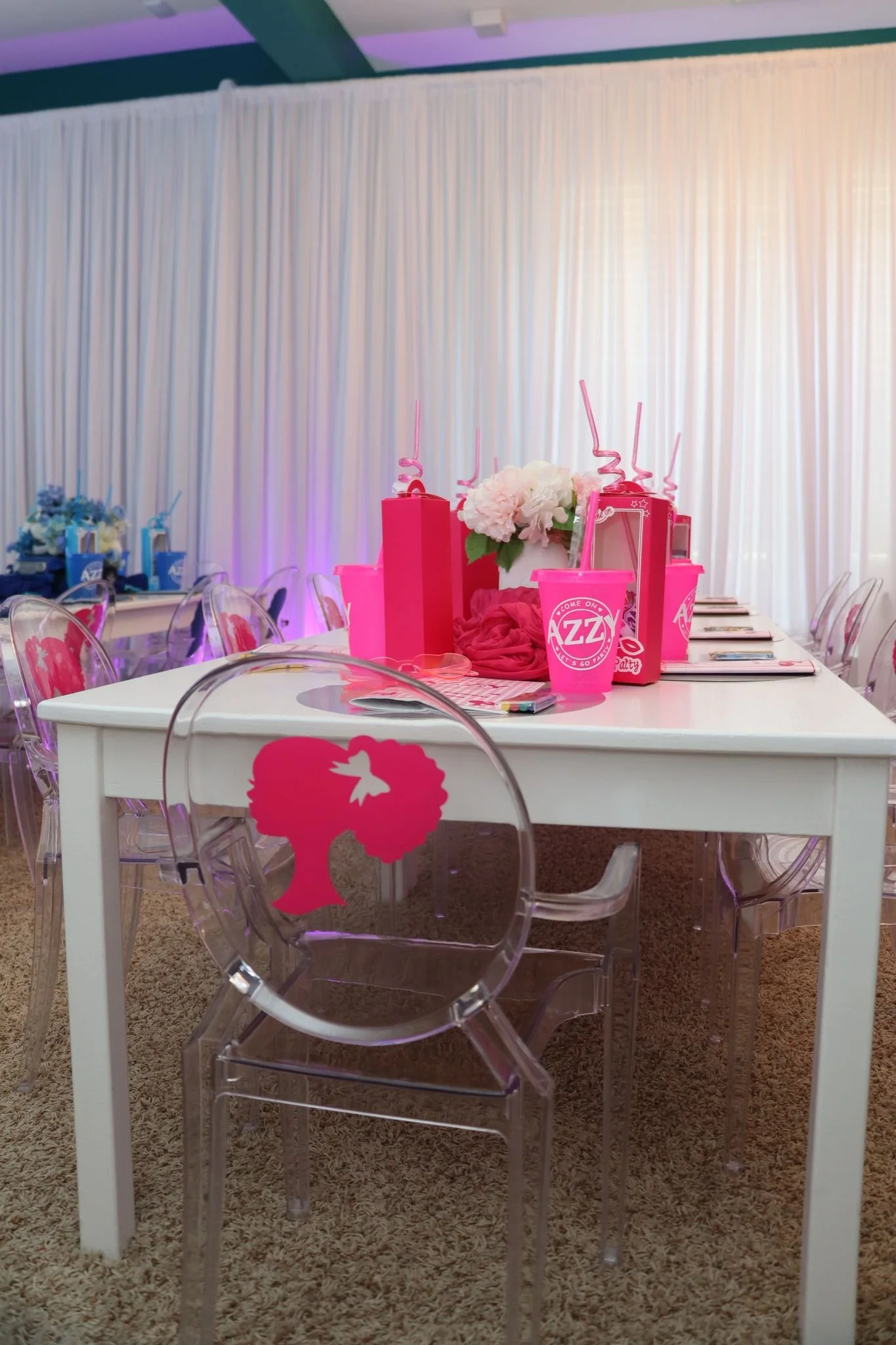Decorated table with pink gift bags, pink cup, flowers, and pink straws, set for a celebration or party. Transparent chairs feature a pink silhouette of a woman with curly hair and a bow. The room has a white backdrop curtain and soft lighting.