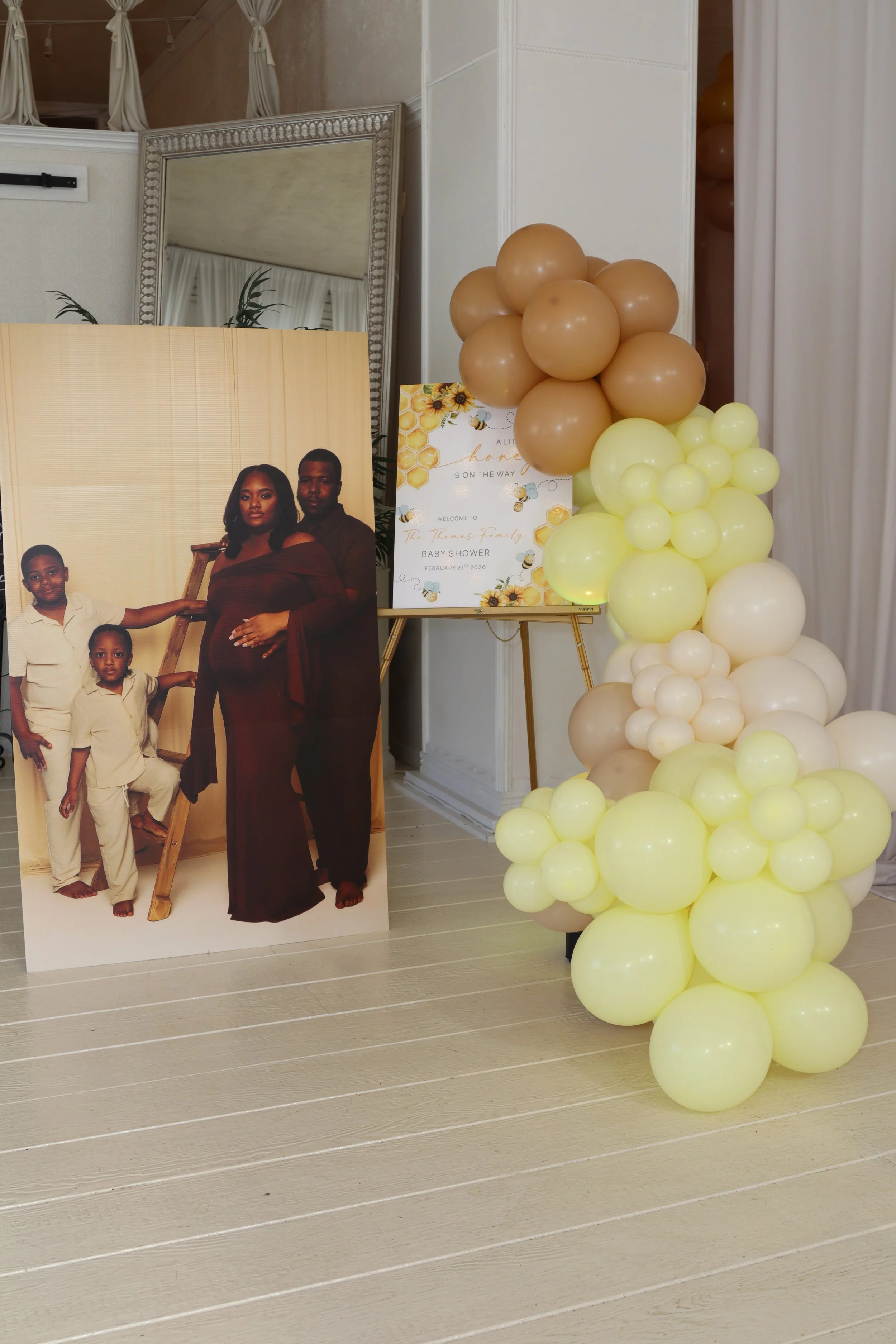 Decorations for a baby shower featuring a family portrait, balloons in beige, yellow, and cream colors, and a sign welcoming guests to the baby shower, date February 21, 2026.