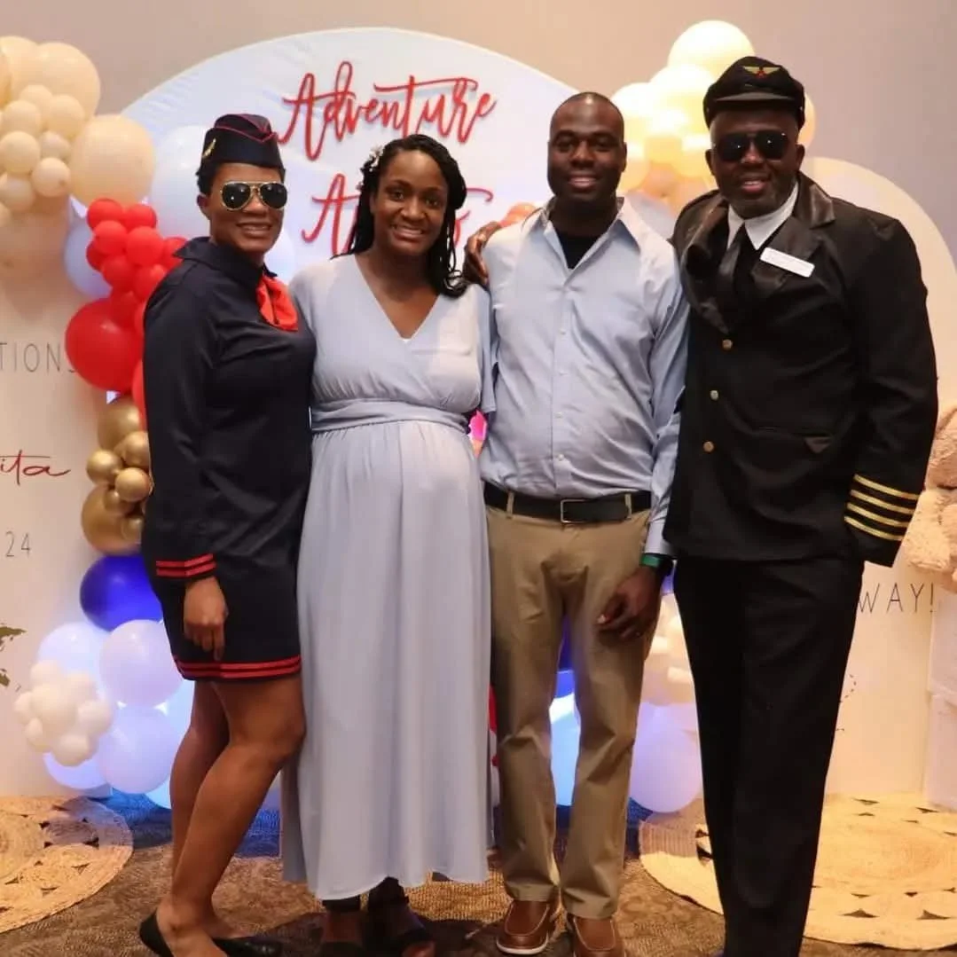 Four people standing together at a celebration with a backdrop that says "Adventure" and "H" in red letters, decorated with balloons in various colors including red, white, gold, and blue.