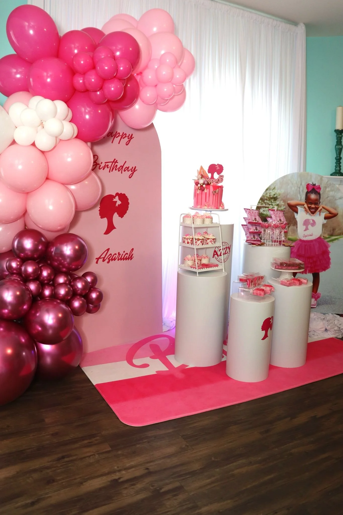 Decorative birthday party setup with pink and white balloons, a birthday cake on a stand, and a girl in pink tutu dress smiling in front of a themed backdrop.